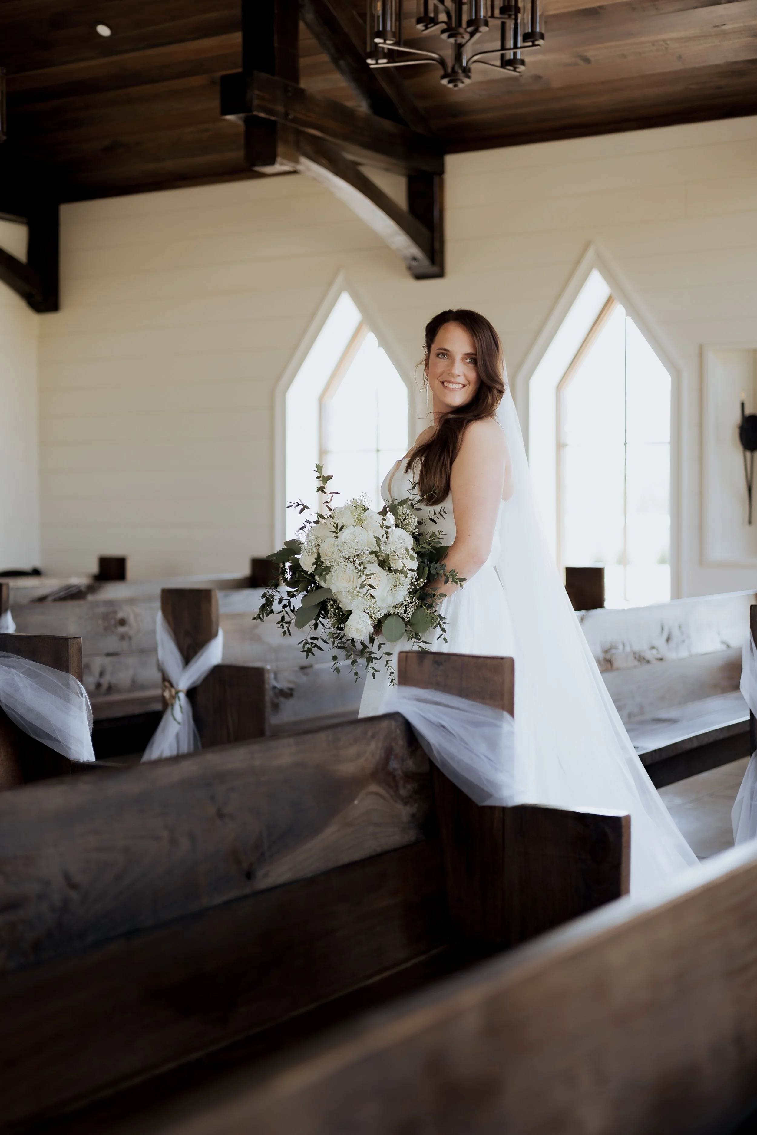 A bride in a white wedding dress holding a bouquet of white flowers inside a wooden chapel with natural light coming through large windows.