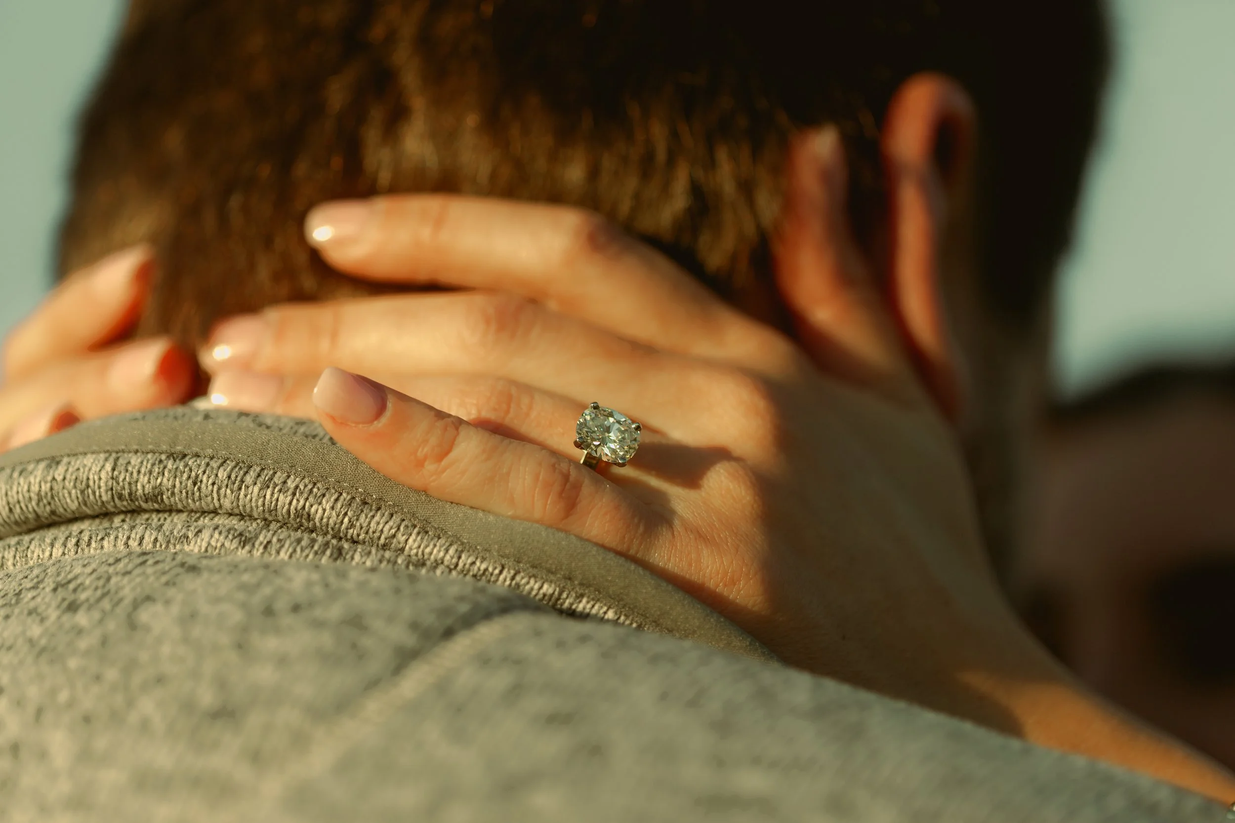 Close-up of a woman with a diamond ring on her ring finger, holding her neck with both hands, with sunlight highlighting her skin and hair.