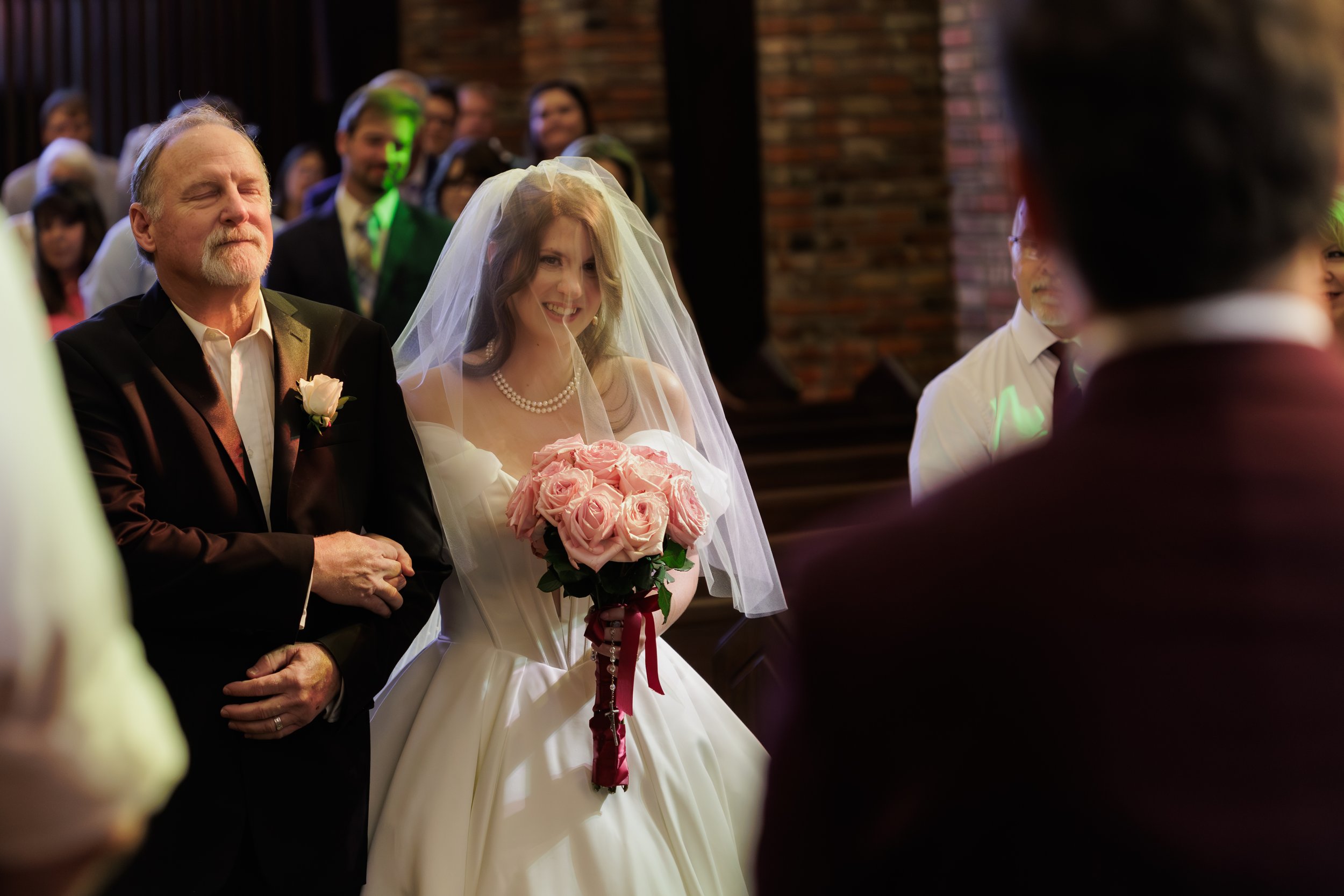 Bride smiling with a bouquet of pink roses, walking down the aisle with her father in a wedding ceremony indoors with guests watching. Chapel of Memories in Starkville, MS on a weding day.