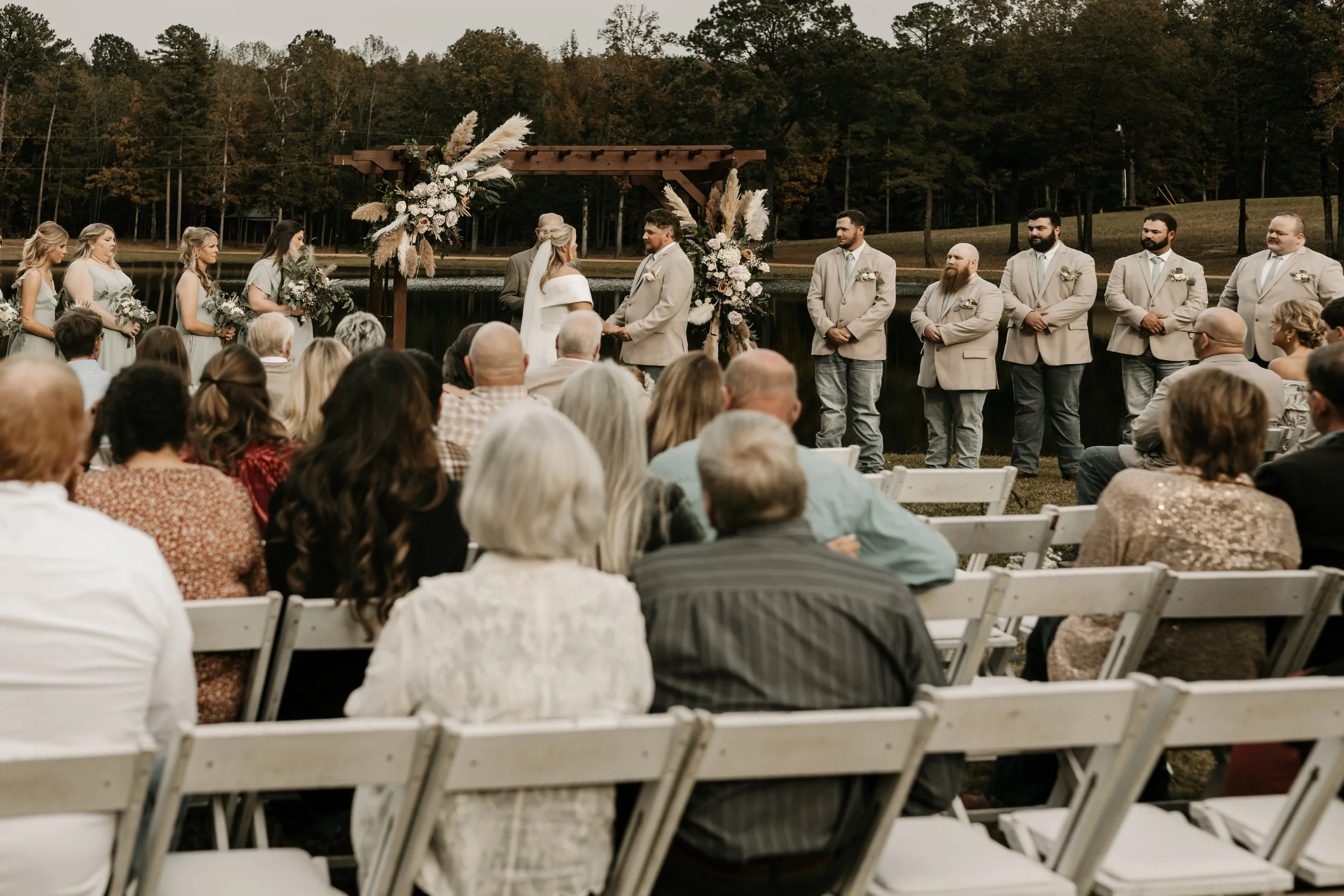 Outdoor wedding ceremony by a lake with the bride and groom exchanging vows, surrounded by bridesmaids and groomsmen, with an audience seated in front.Behind The Pines Wedding Venue. Wedding Photography in Mississippi.