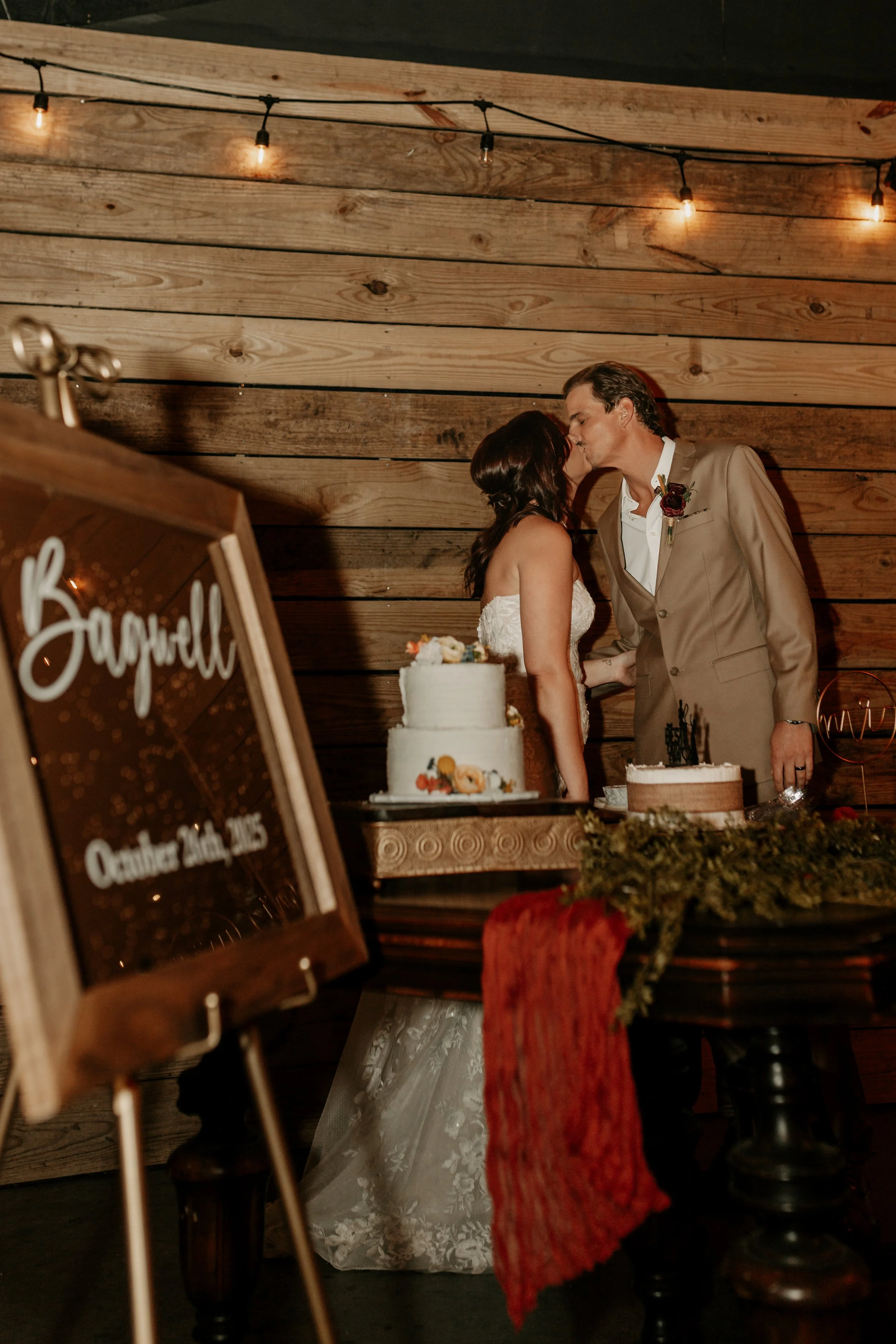 A newlywed couple sharing a kiss at their wedding reception, standing behind a table with wedding cakes, in a rustic wooden setting with string lights overhead. The Stables in Smithville, MS. Wedding photography in Mississippi.