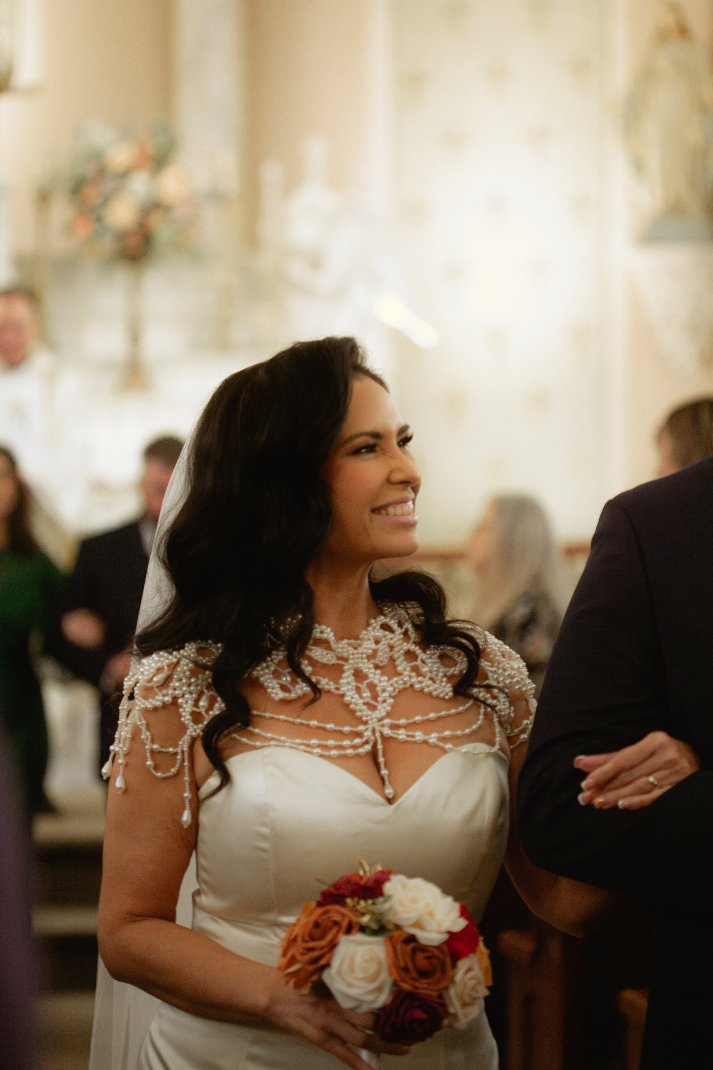 A woman in a white wedding dress with a pearl necklace and a veil, smiling and holding a bouquet of roses, attending a wedding ceremony.