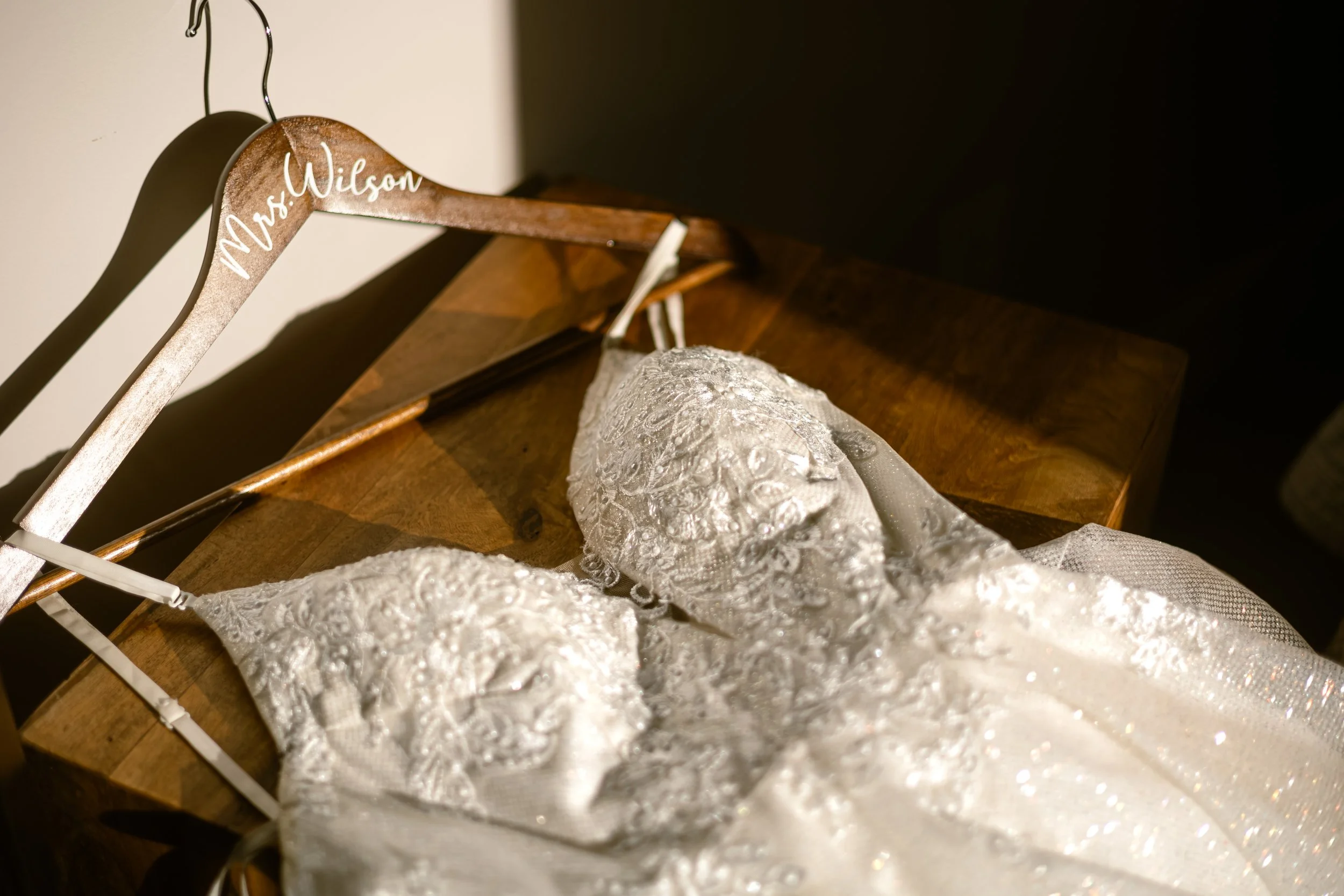 A wedding dress with intricate lace and beading detail resting on a wooden surface, hanging on a wooden hanger with "Mrs. Wilson" written on it.