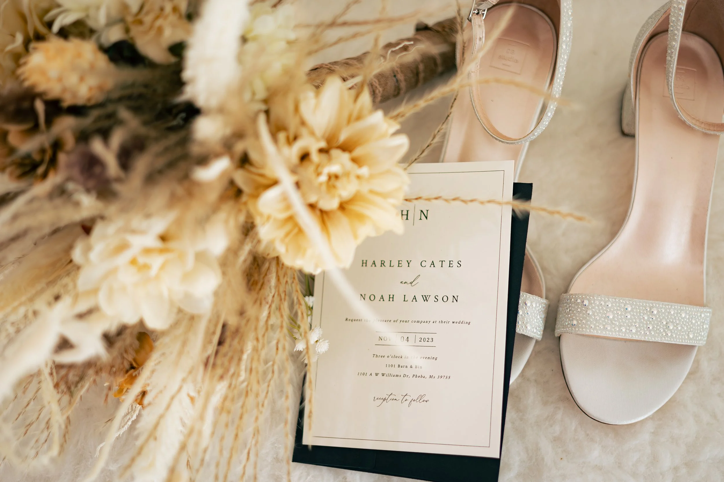 A wedding invitation card, placed beside a pair of white, decorated high heel shoes with embellished straps, with dried flowers in the foreground.