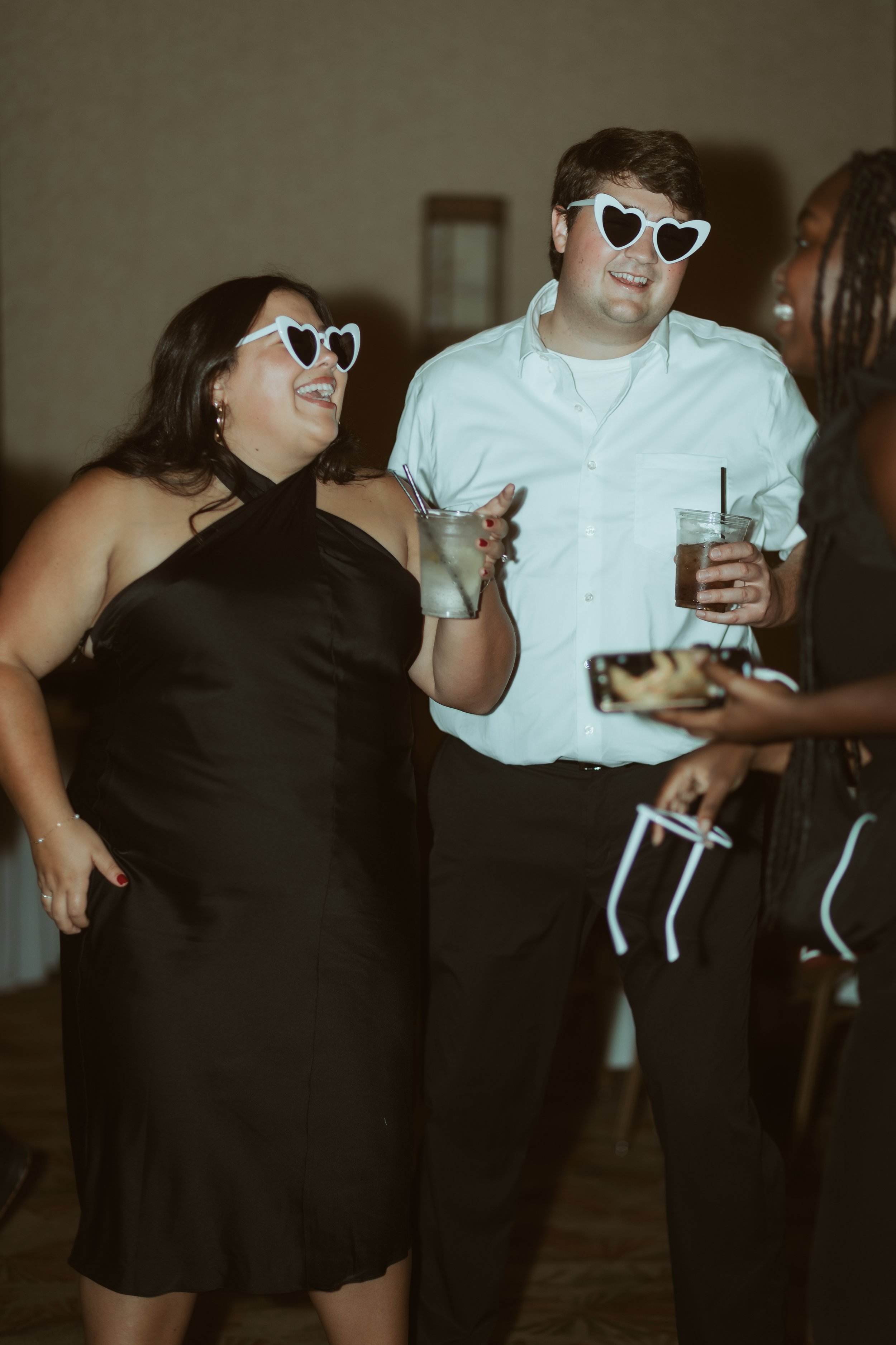Three friends wearing heart-shaped sunglasses enjoying drinks and laughing at a social gathering.