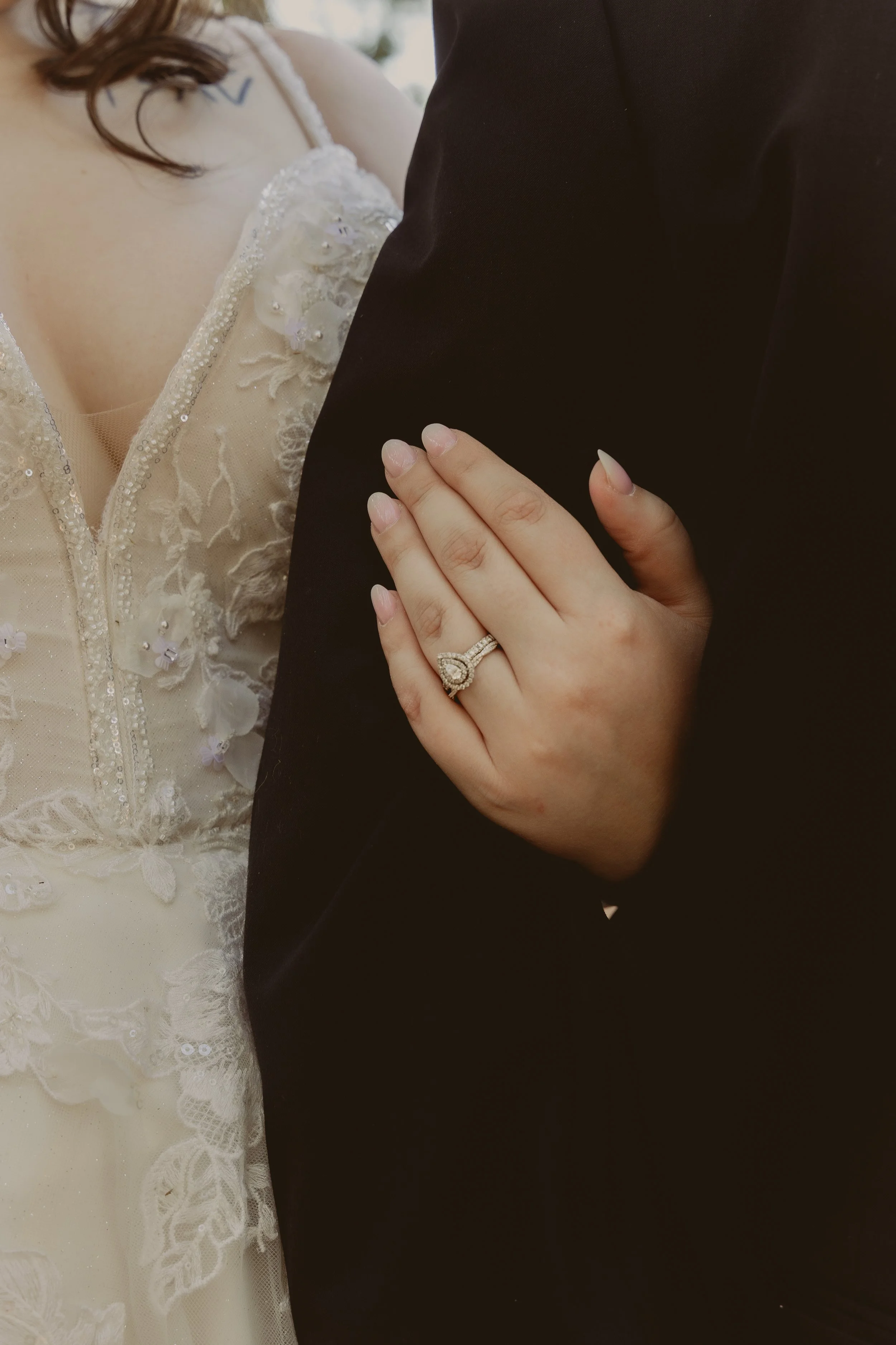 Close-up of a wedding ring on a woman's finger, with her hand resting on a man's arm dressed in a black suit, and part of a white bridal gown with lace and beadwork visible. Wedding Photography in Starkville, MS.