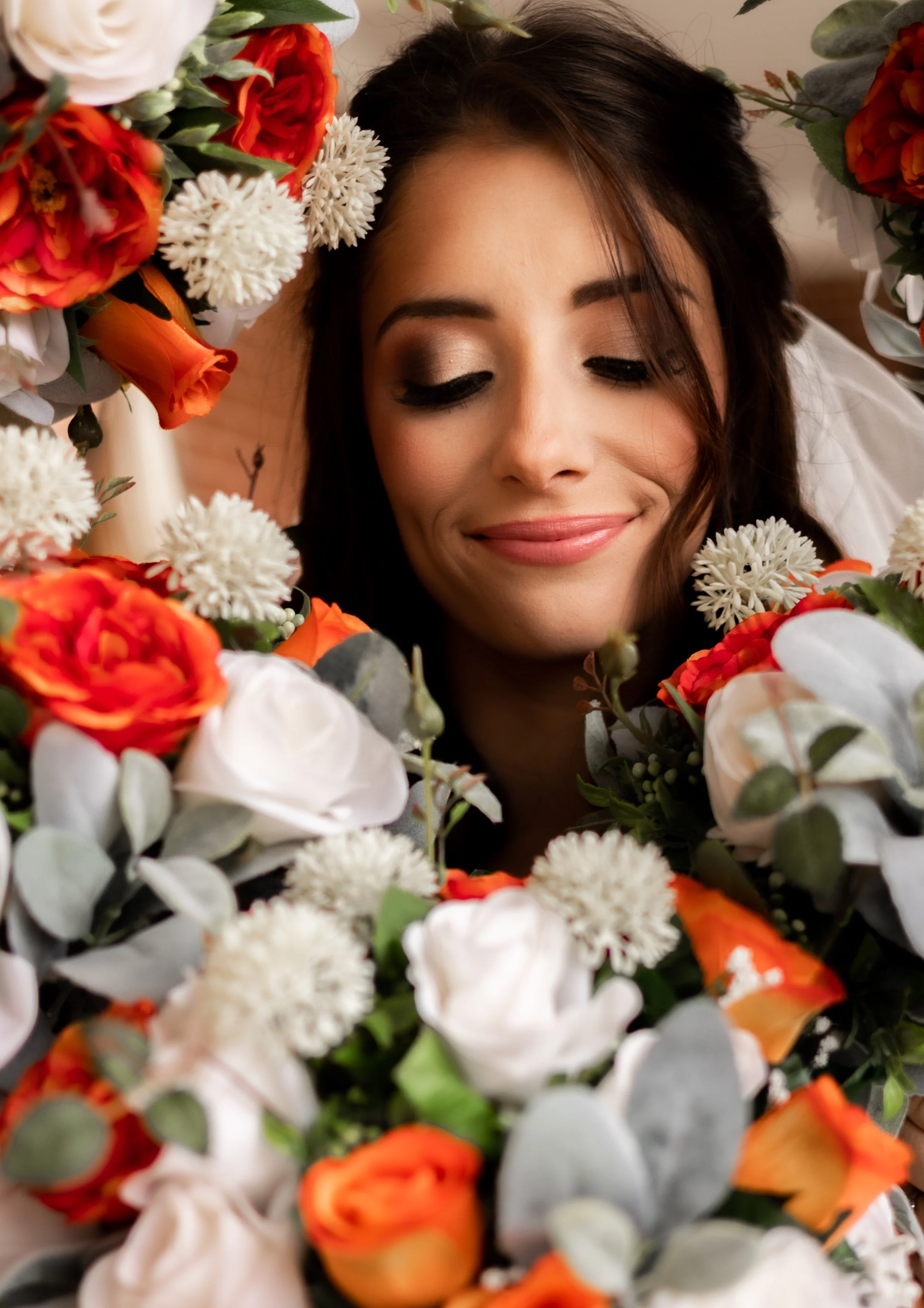 A woman with makeup and dark hair, smiling peacefully, surrounded by an assortment of orange, white, and peach-colored flowers, including roses, ranunculus, and small white blossoms.