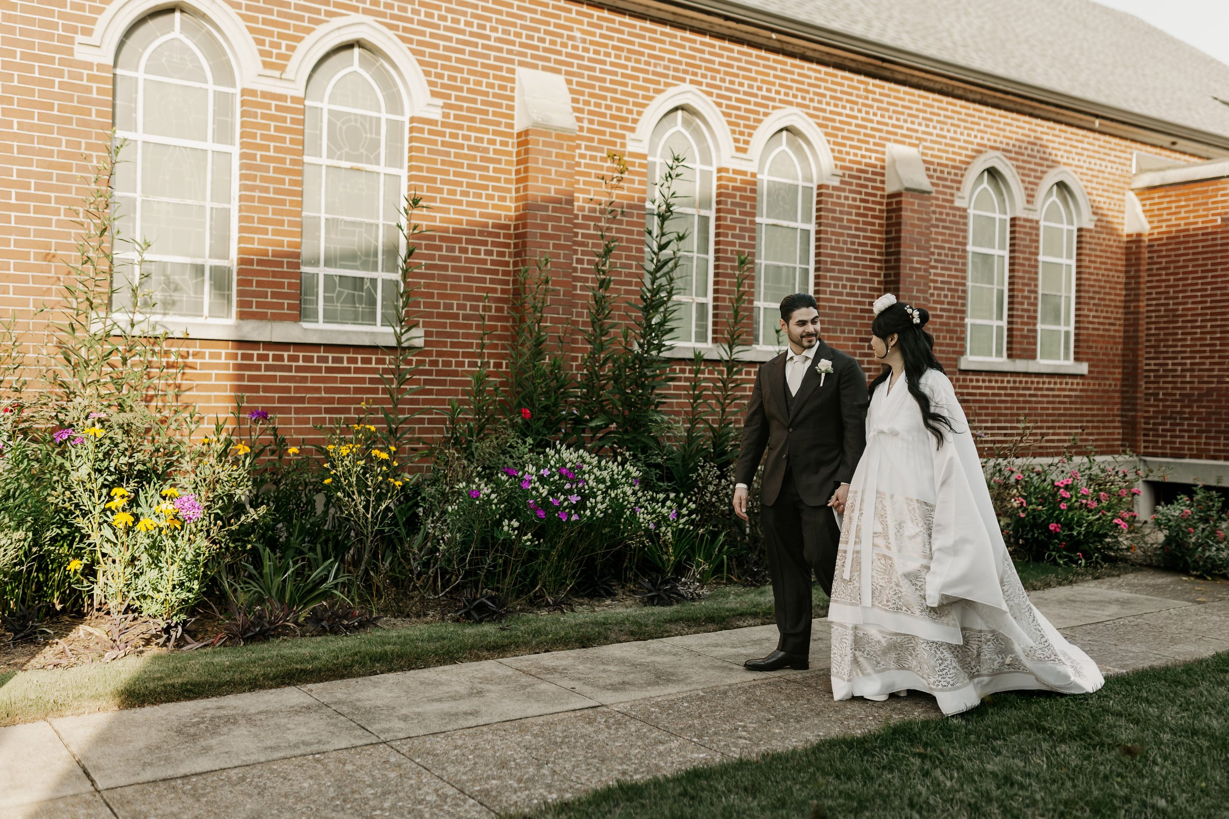 A bride and groom holding hands and walking outside a brick church with arched windows, surrounded by colorful flowers.