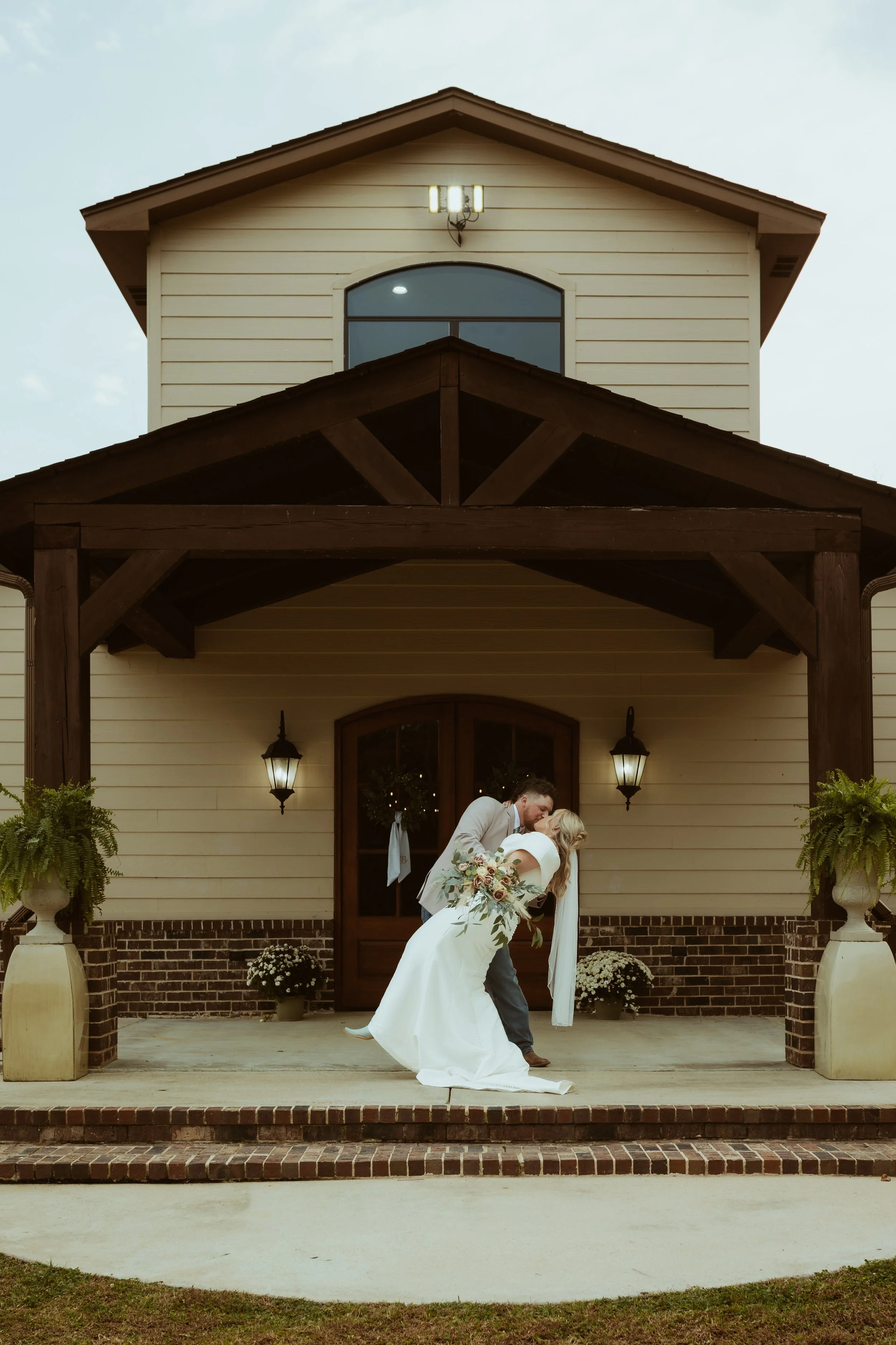 A bride and groom sharing a kiss on the porch of a house during a wedding photo. The bride is in a white wedding dress holding a bouquet, and the groom is in a light-colored suit. The house has a beige exterior with dark wooden accents, two wall-moun