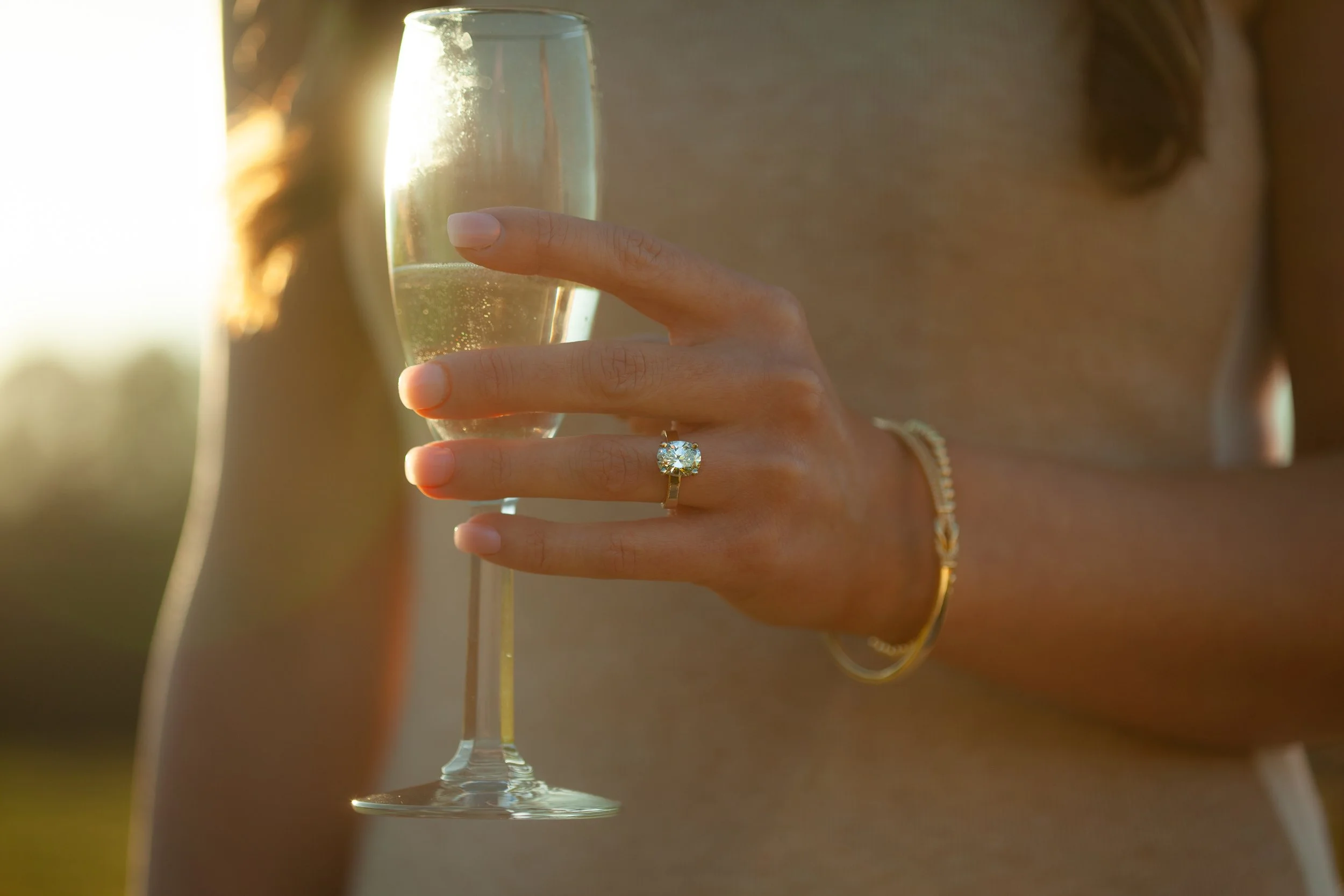 A woman holding a glass of champagne with an engagement ring on her finger, while wearing a gold bracelet