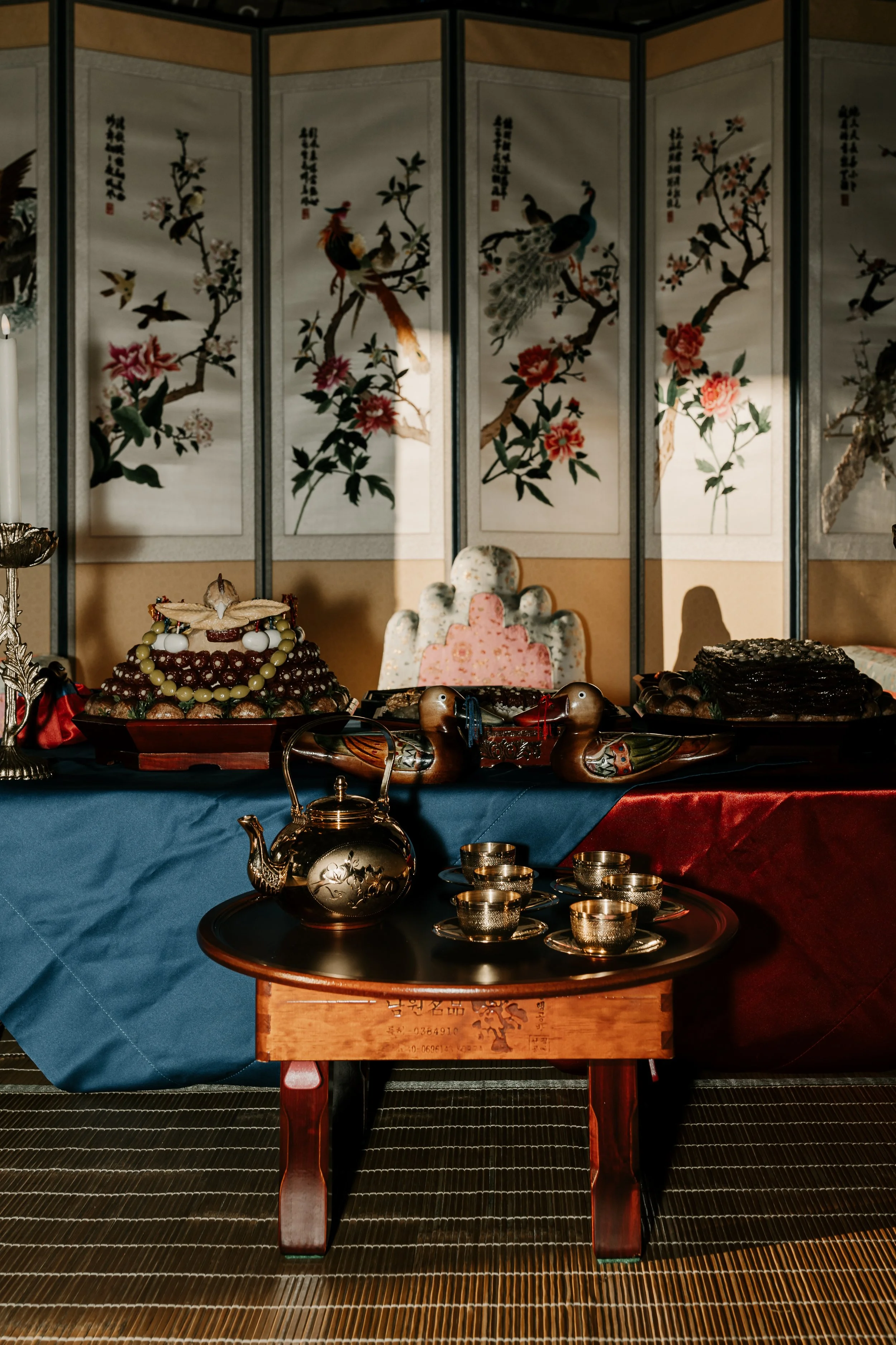 Bohemian-style wedding or celebration table with traditional Korean foods, tea set, decorative ducks, and a folding screen with floral and bird paintings in the background.