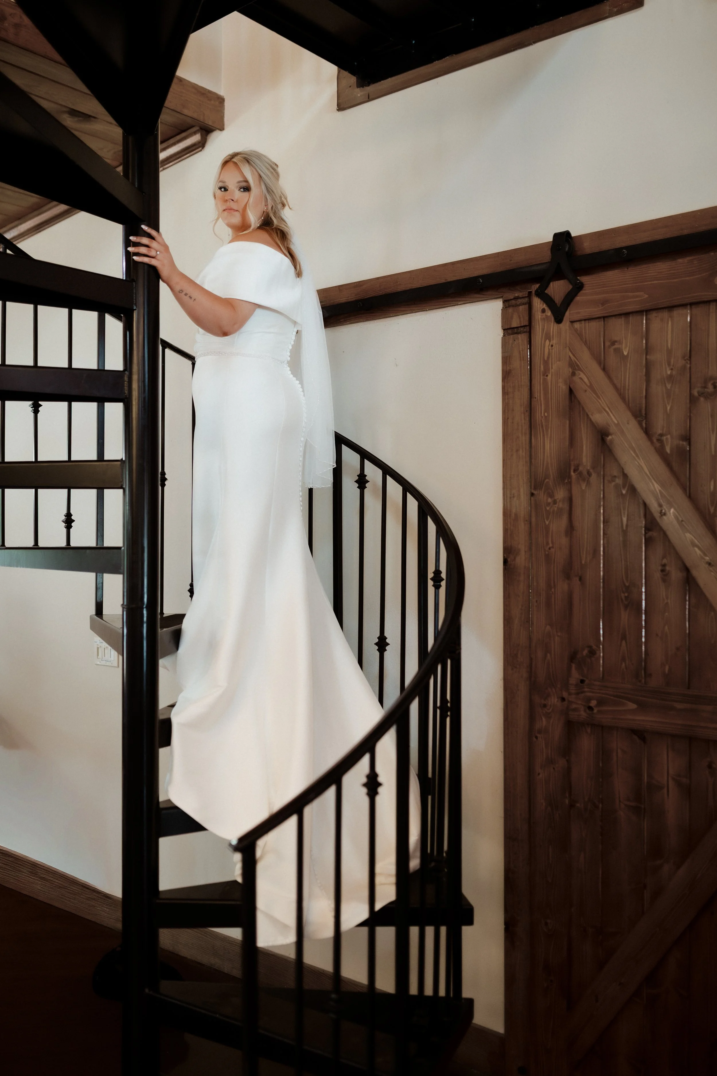 Bride in a white wedding dress and veil standing on a black spiral staircase inside a rustic wooden building. Behind The Pines Wedding Venue. Wedding Photography in Mississippi.