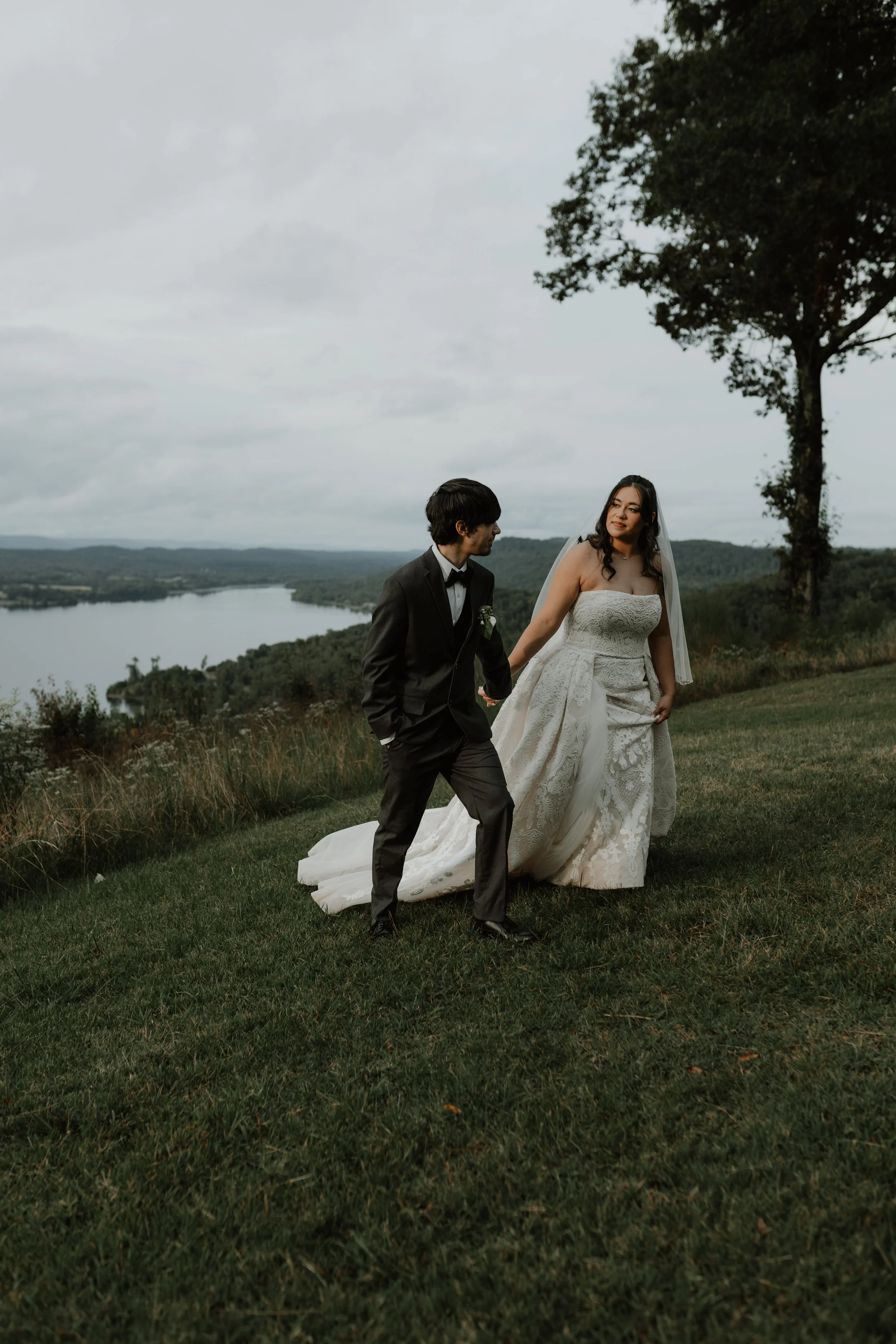 A bride and groom in wedding attire holding hands outdoors on a grassy hillside with a river and landscape in the background.