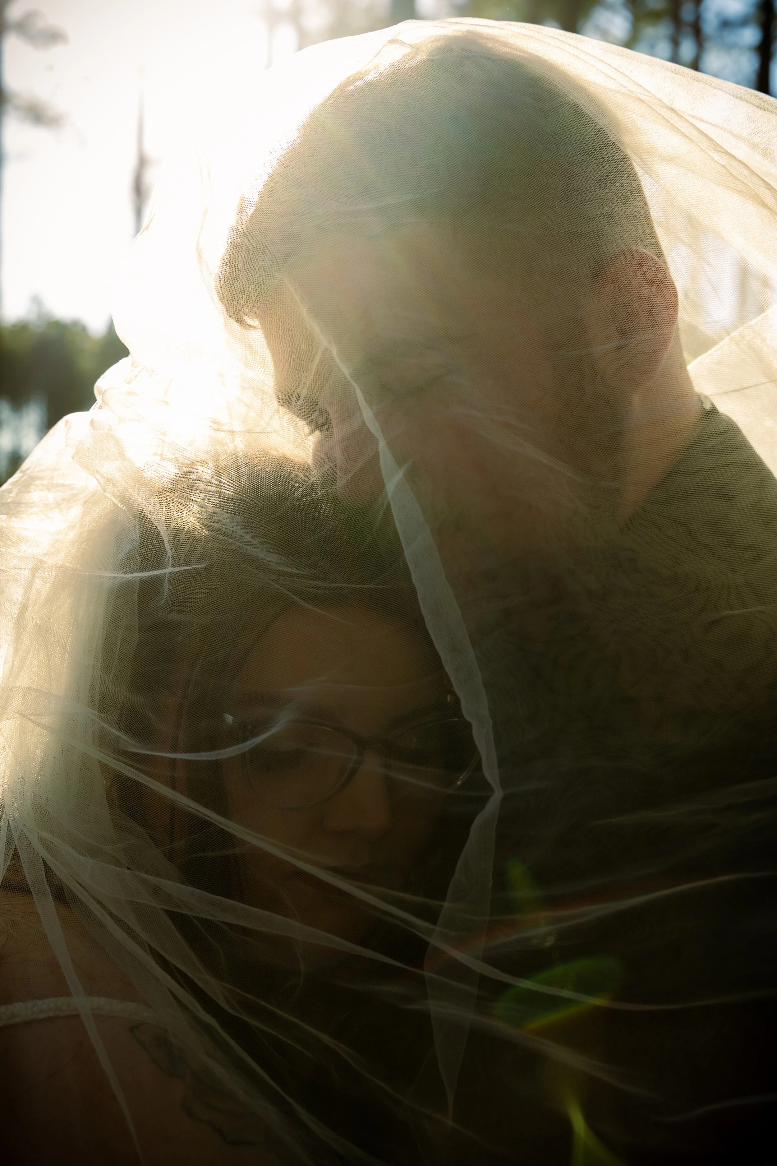 A couple under a white veil, backlit by sunlight, outdoors, with trees in the background.