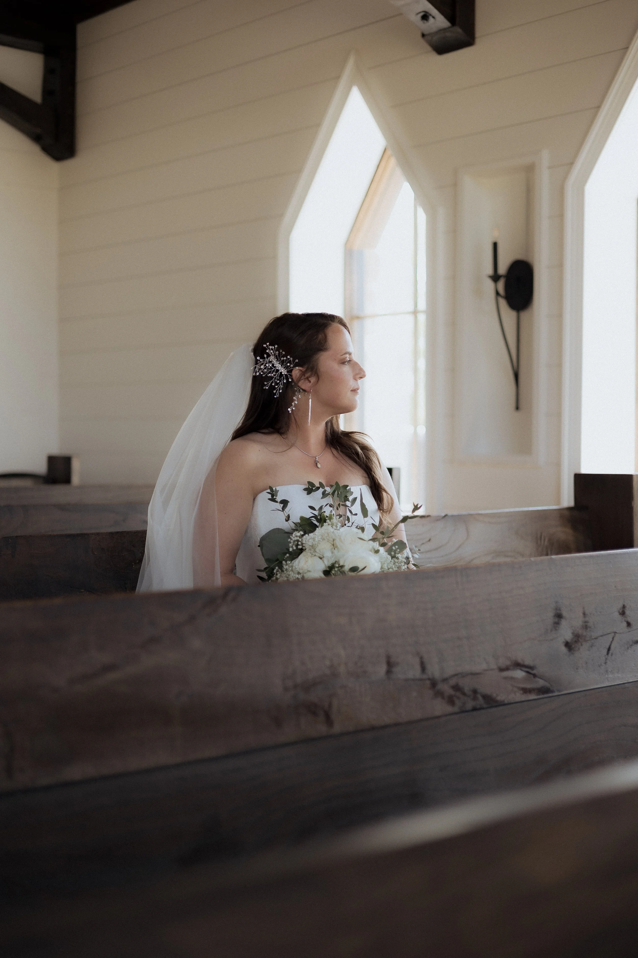 A bride sitting indoors, holding a bouquet of white flowers and greenery, wearing a strapless wedding dress and a veil, with her hair styled with a decorative hairpiece. She is looking to the right, with soft natural light coming through windows with