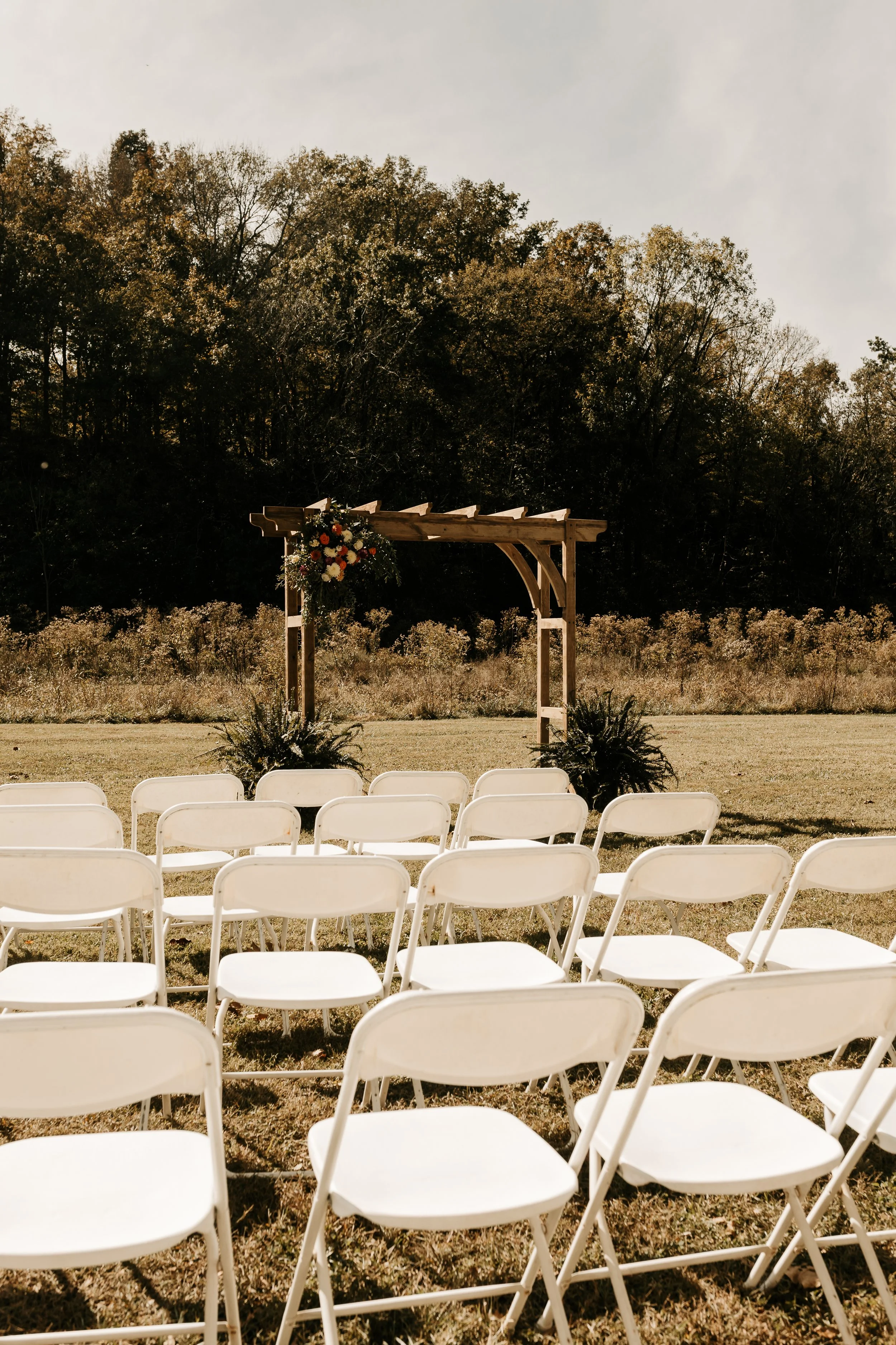 Outdoor wedding ceremony setup with white folding chairs arranged on grass facing a wooden arch decorated with flowers and greenery, set in a natural outdoor setting with trees in the background. Alabama Wedding Photography.