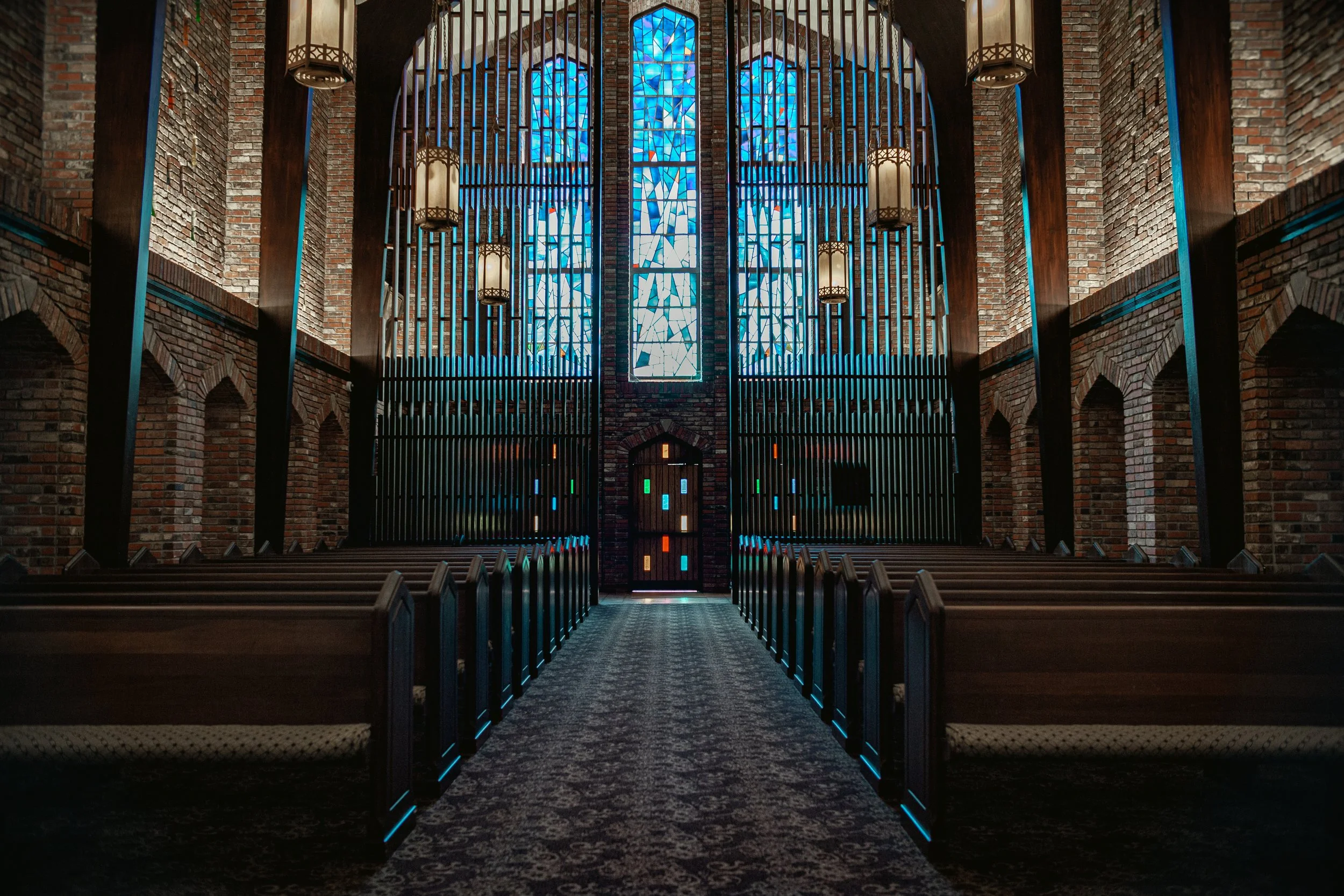 Interior of a church with tall blue stained glass windows, brick walls, wooden pews, and hanging lamps. Chapel of Memories in Starkville, MS on a weding day.