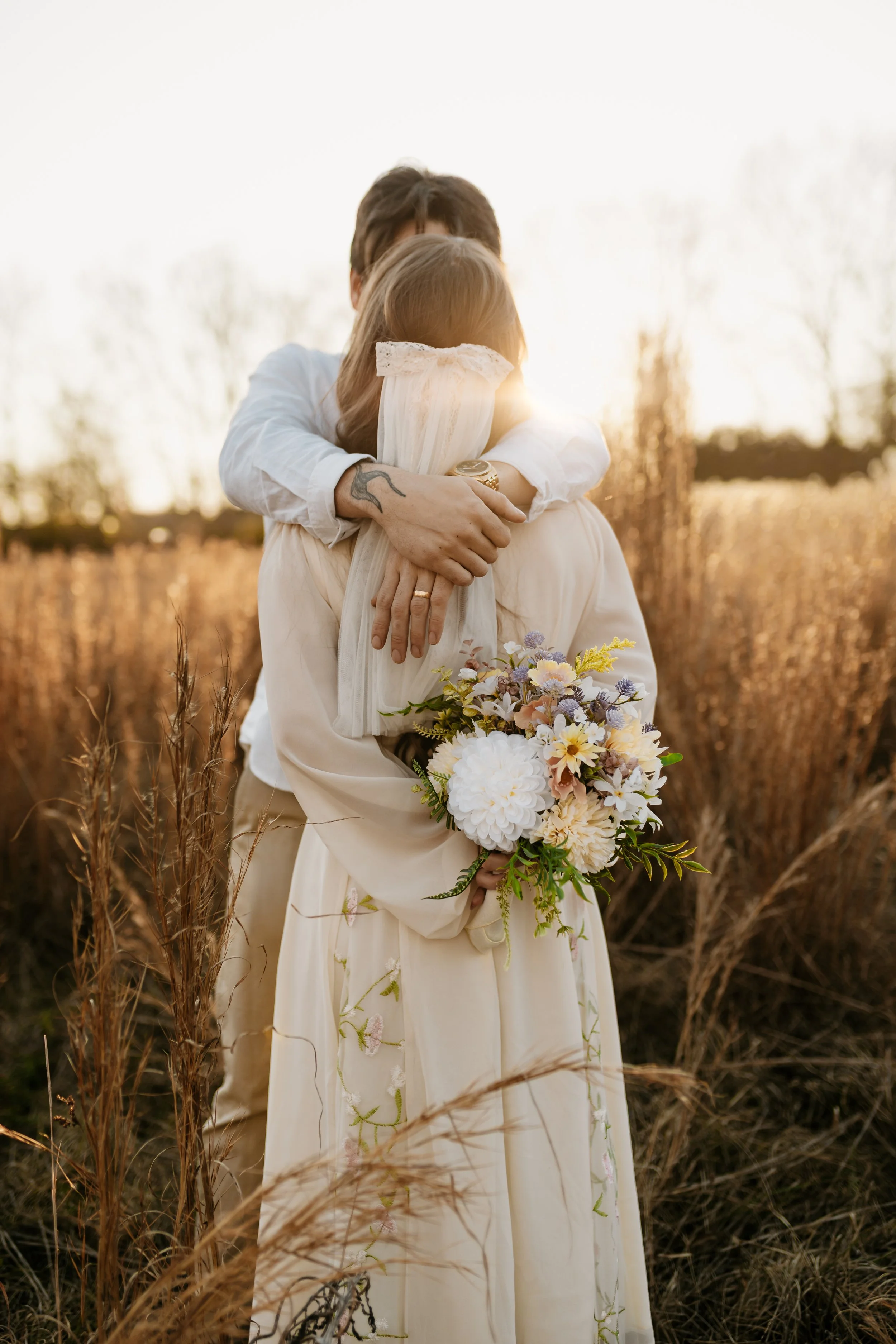 A newlywed couple standing on a grassy area next to a pond, with lush green trees in the background, smiling and embracing.