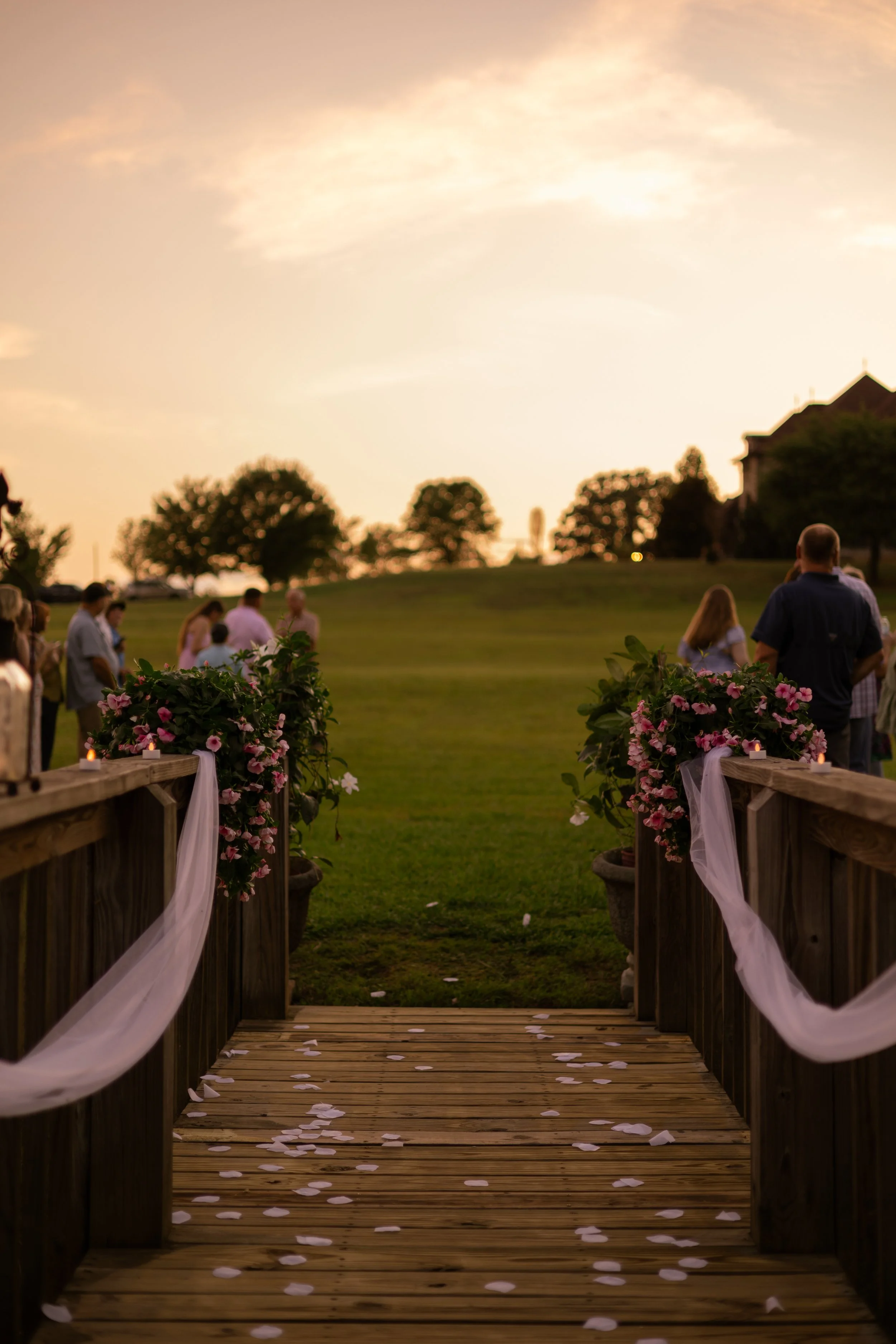 A wedding aisle decorated with pink flowers and white tulle on wooden railings, scattered with white rose petals, leading to an outdoor ceremony site during sunset with guests gathered and trees in the distance.