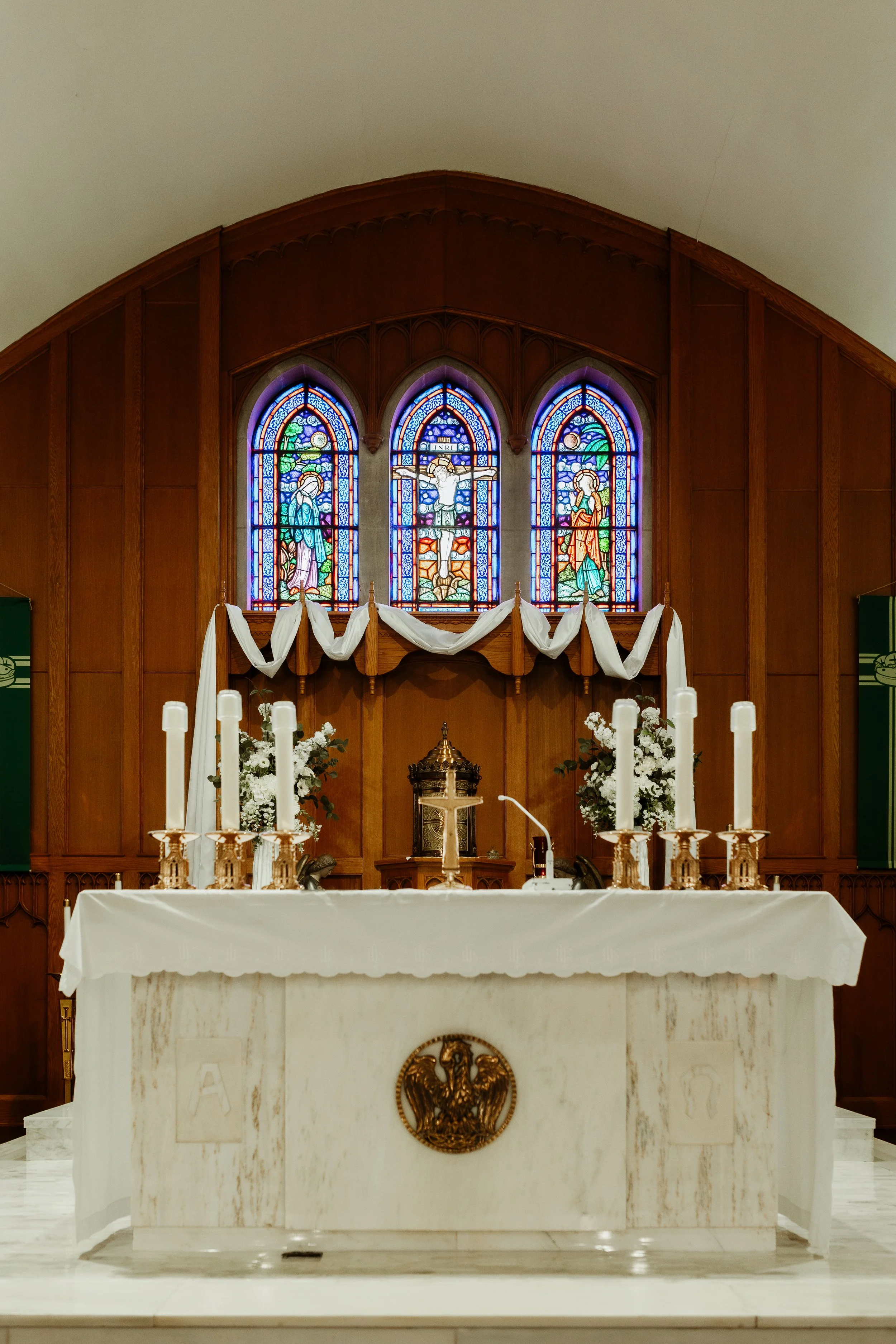 Inside a church with a white altar decorated with candles and a crucifix, and stained glass windows depicting religious figures