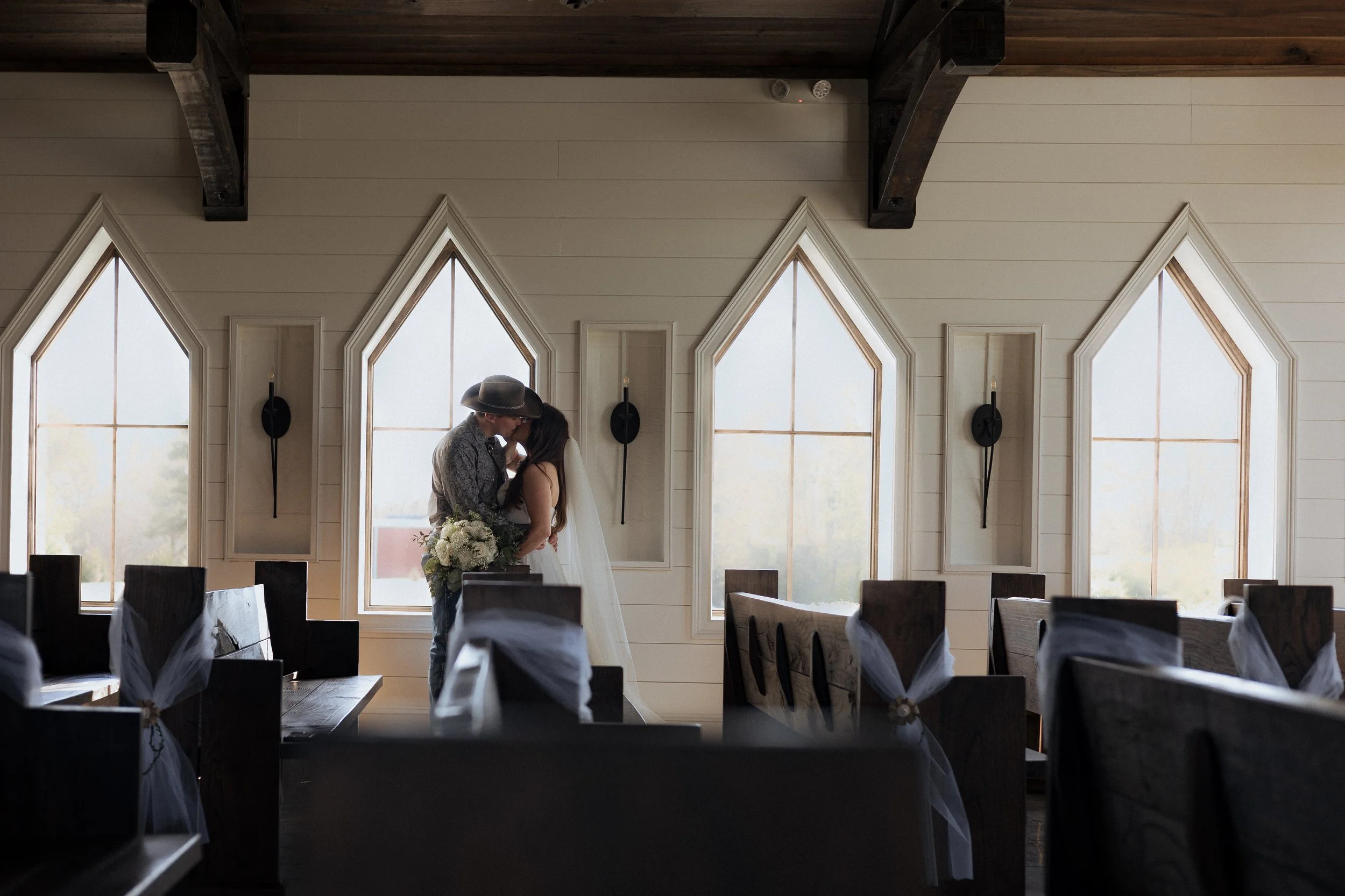 A couple sharing a kiss at an altar in a church during their wedding. The bride is holding a bouquet of white flowers, and the groom is wearing a cowboy hat. The church has large, pointed windows with natural light streaming in, and decorated pews wi