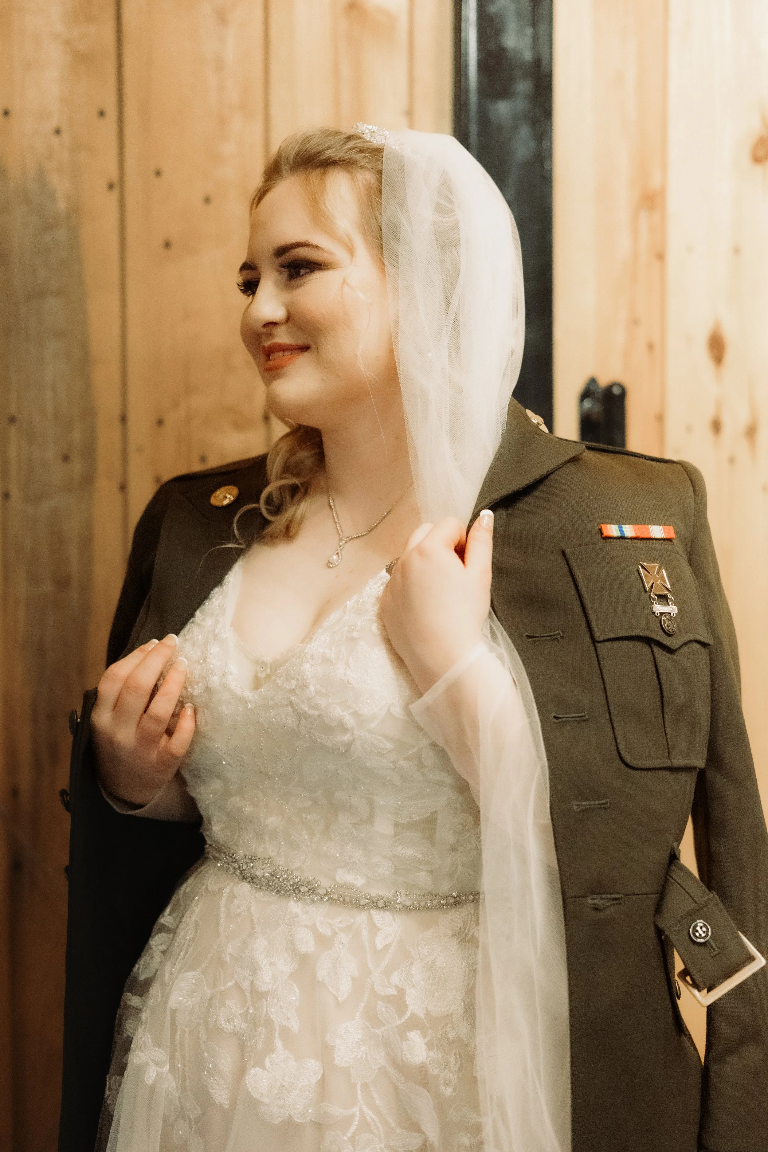 A bride in a wedding dress and veil wearing a military uniform, standing in front of a wooden wall. Military bride wedding photography inspiration.