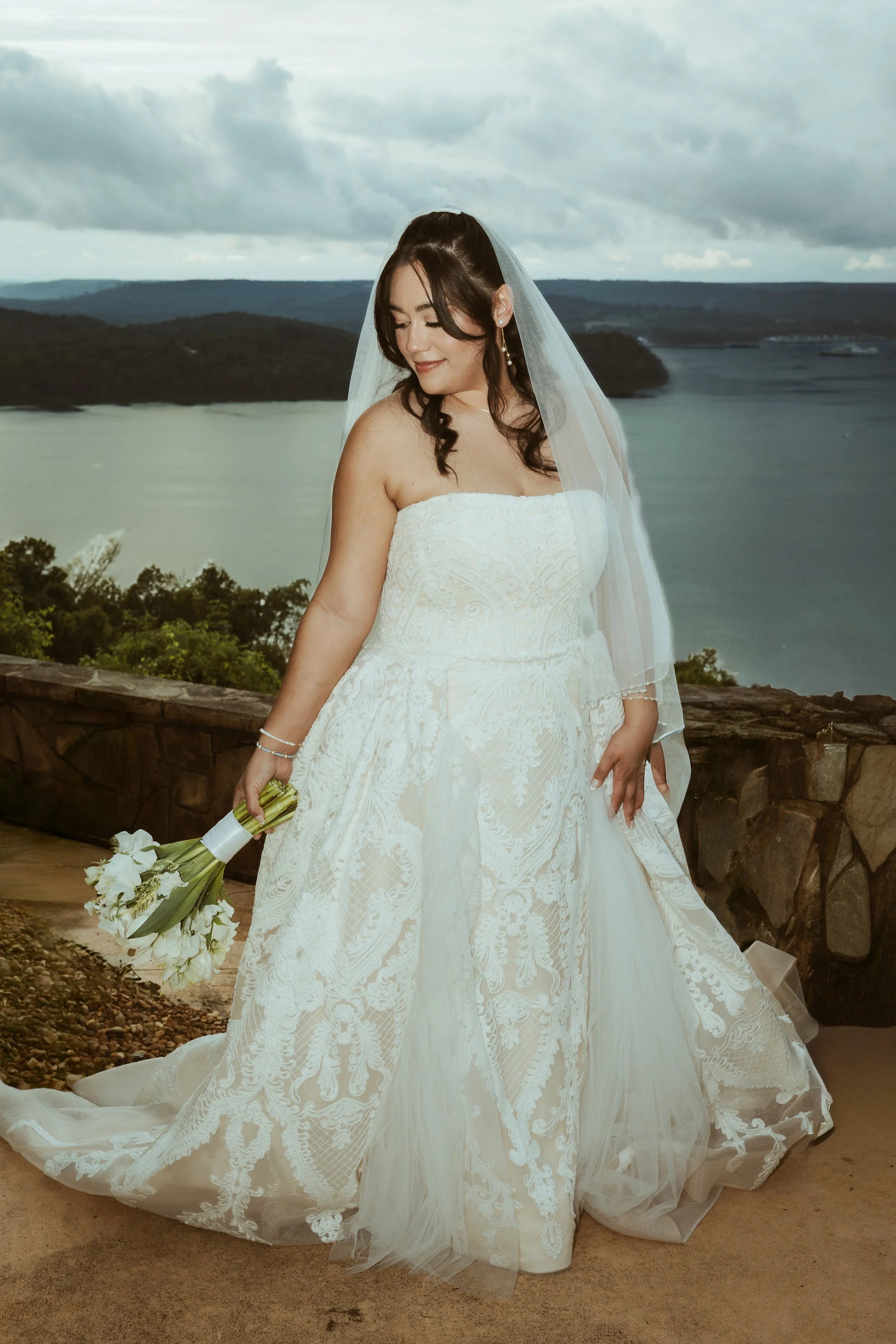 Bride standing outdoors holding a bouquet of white flowers, wearing a strapless white lace wedding gown and veil, with a scenic lake and cloudy sky in the background.
