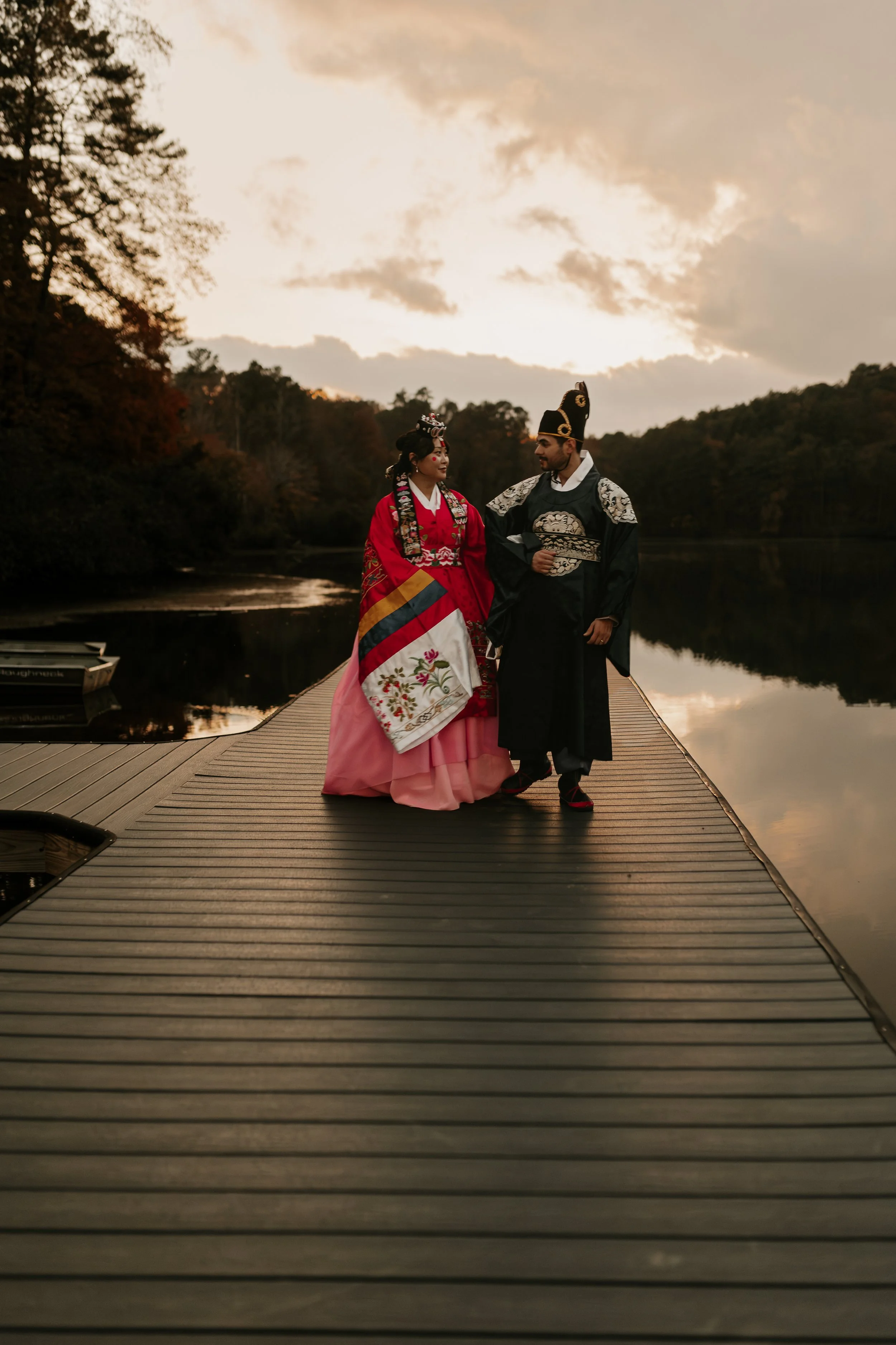 A couple dressed in traditional Korean wedding attire stands on a wooden dock beside a lake during sunset. Birmingham, AL wedding photography.