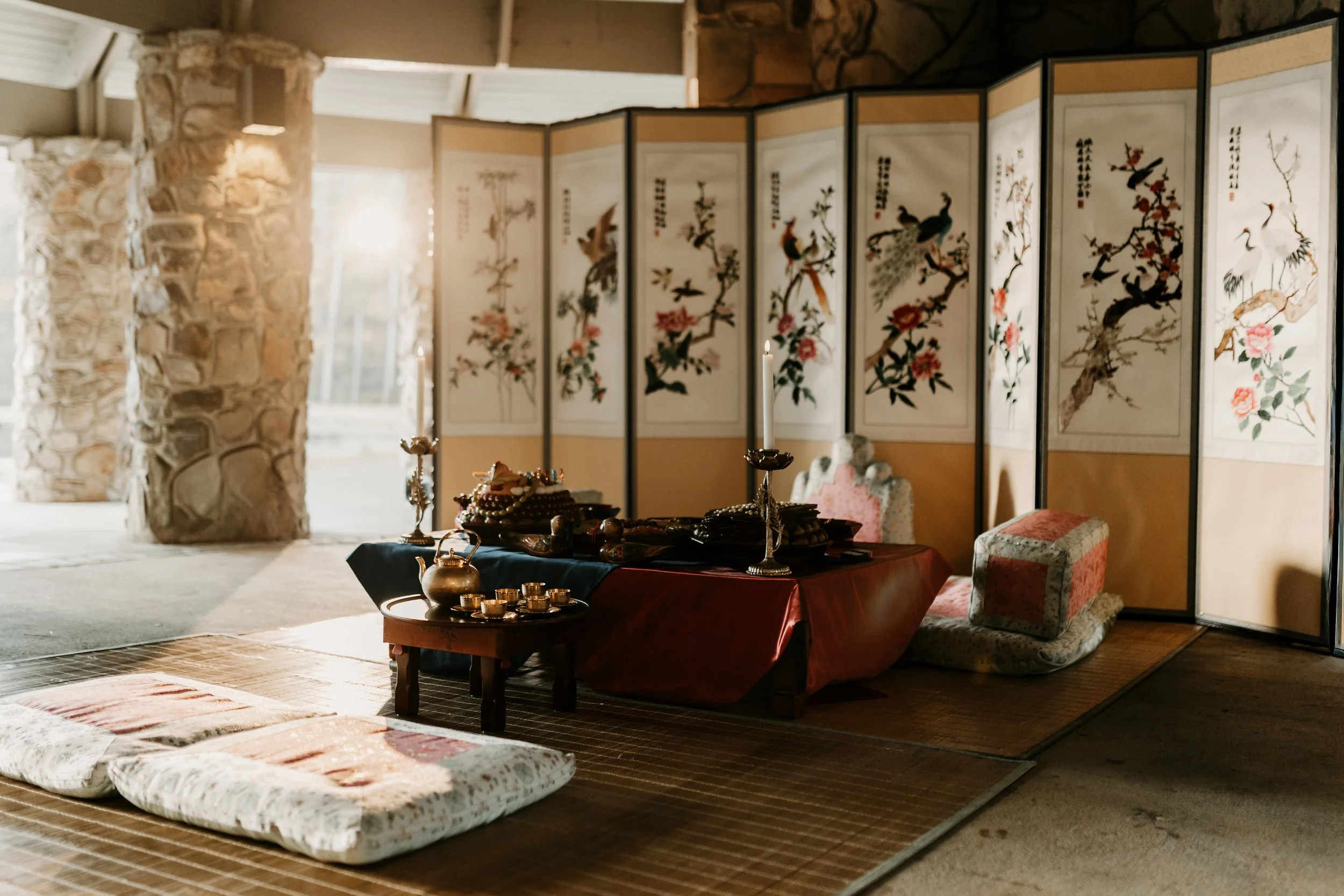 A traditional Japanese indoor scene with a low table covered in red cloth, decorated with candles, teapot, and dishes. Surrounding the table are floor cushions and a folding screen with birds and floral paintings. The setting features stone pillars a