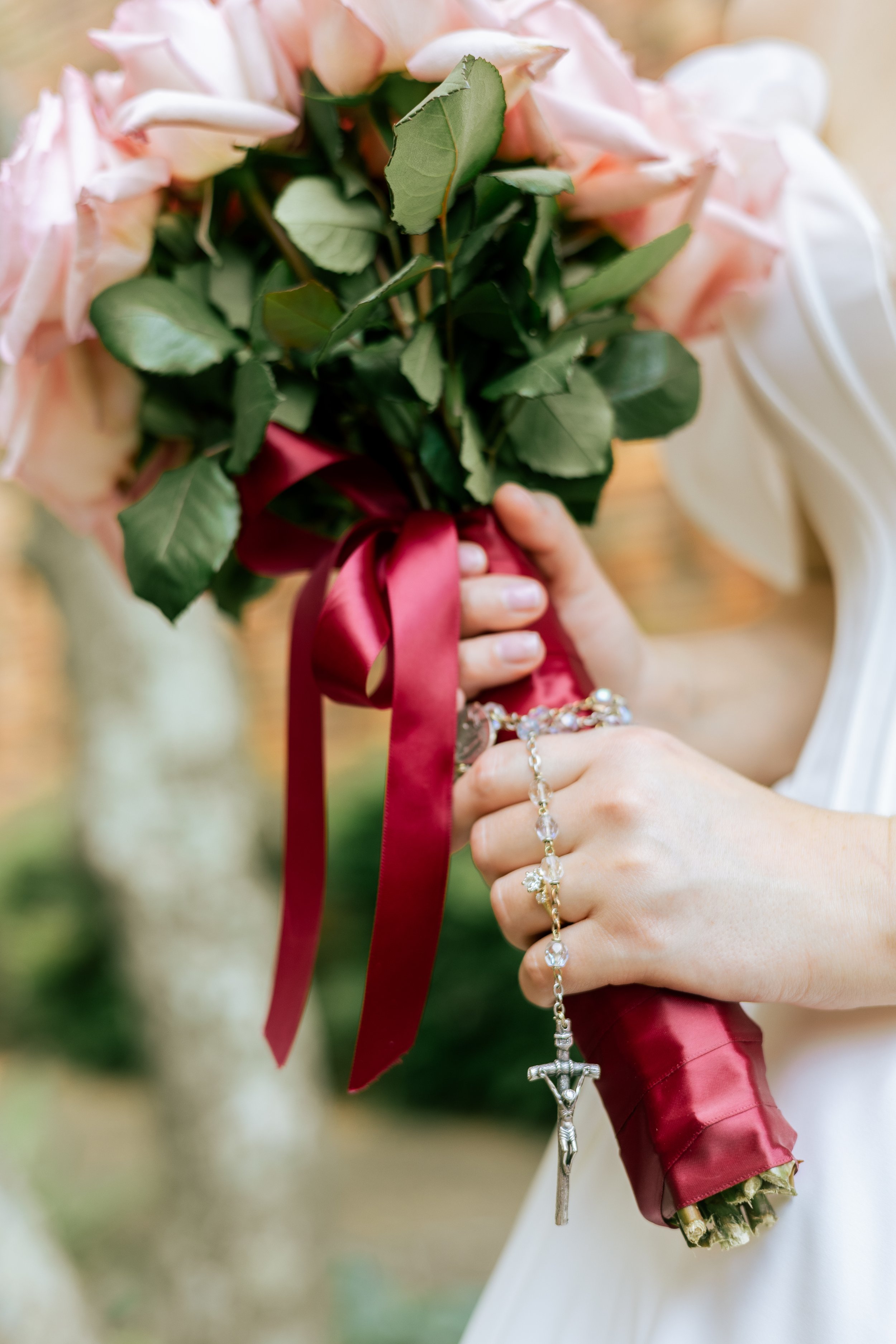 A person holding a bouquet of pink roses tied with a red ribbon, with a Catholic rosary hanging from their hand.