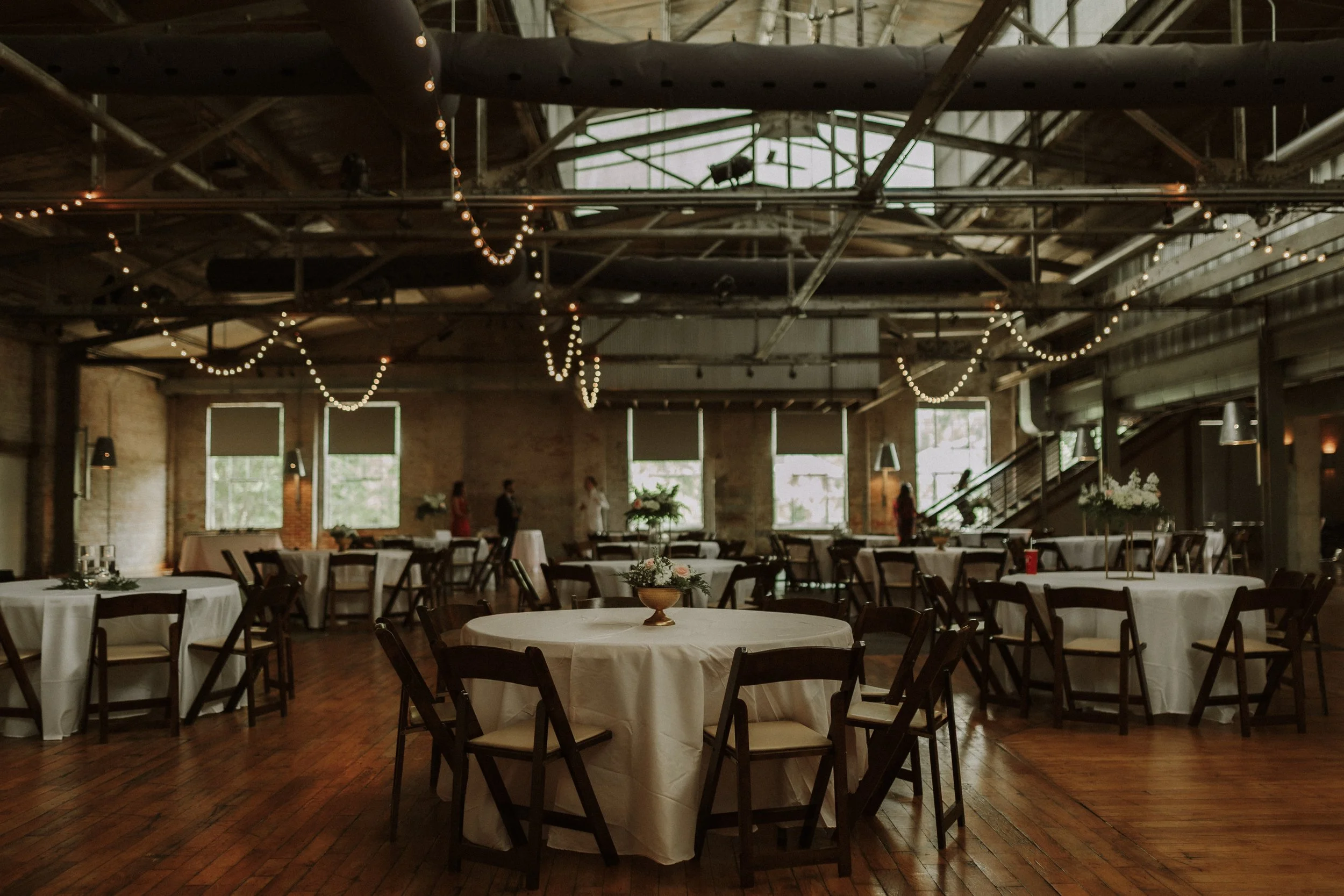 An empty event space with round tables covered in white tablecloths and decorated with floral centerpieces, surrounded by dark wooden chairs. String lights hang from the ceiling, and large windows let in natural light. A staircase is visible on the r