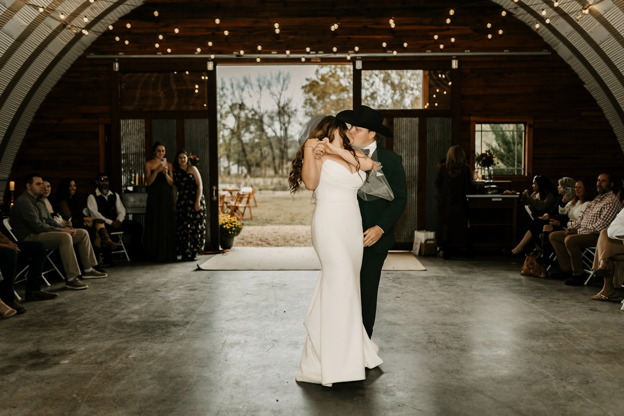 Couple dances closely indoors during a wedding reception with seated guests watching, natural light, rustic venue, and string lights overhead.