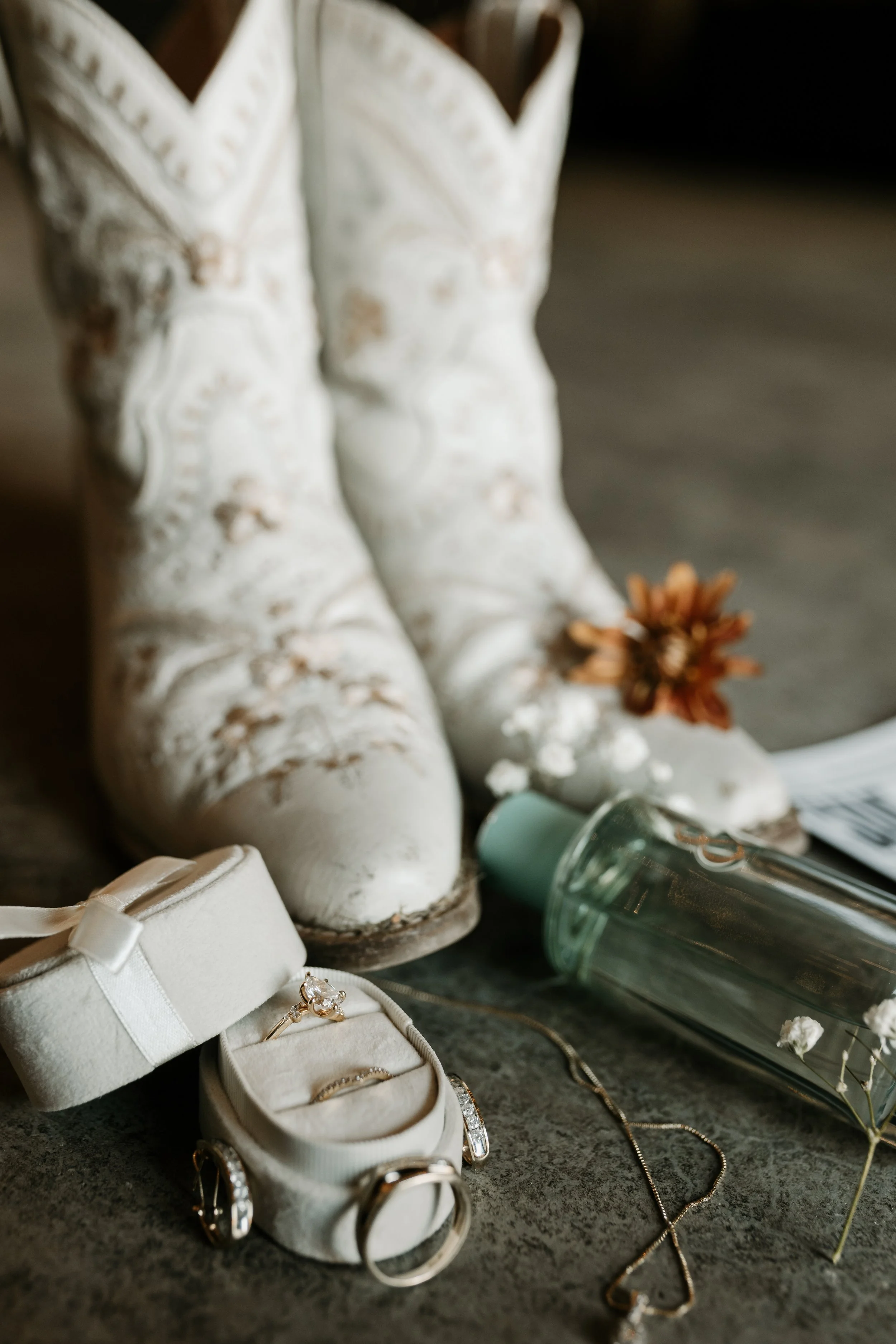 Vintage white cowboy boots, jewelry including rings and necklaces, a small box, a flower, and a water bottle on a dark surface. Unique detail wedding photography.