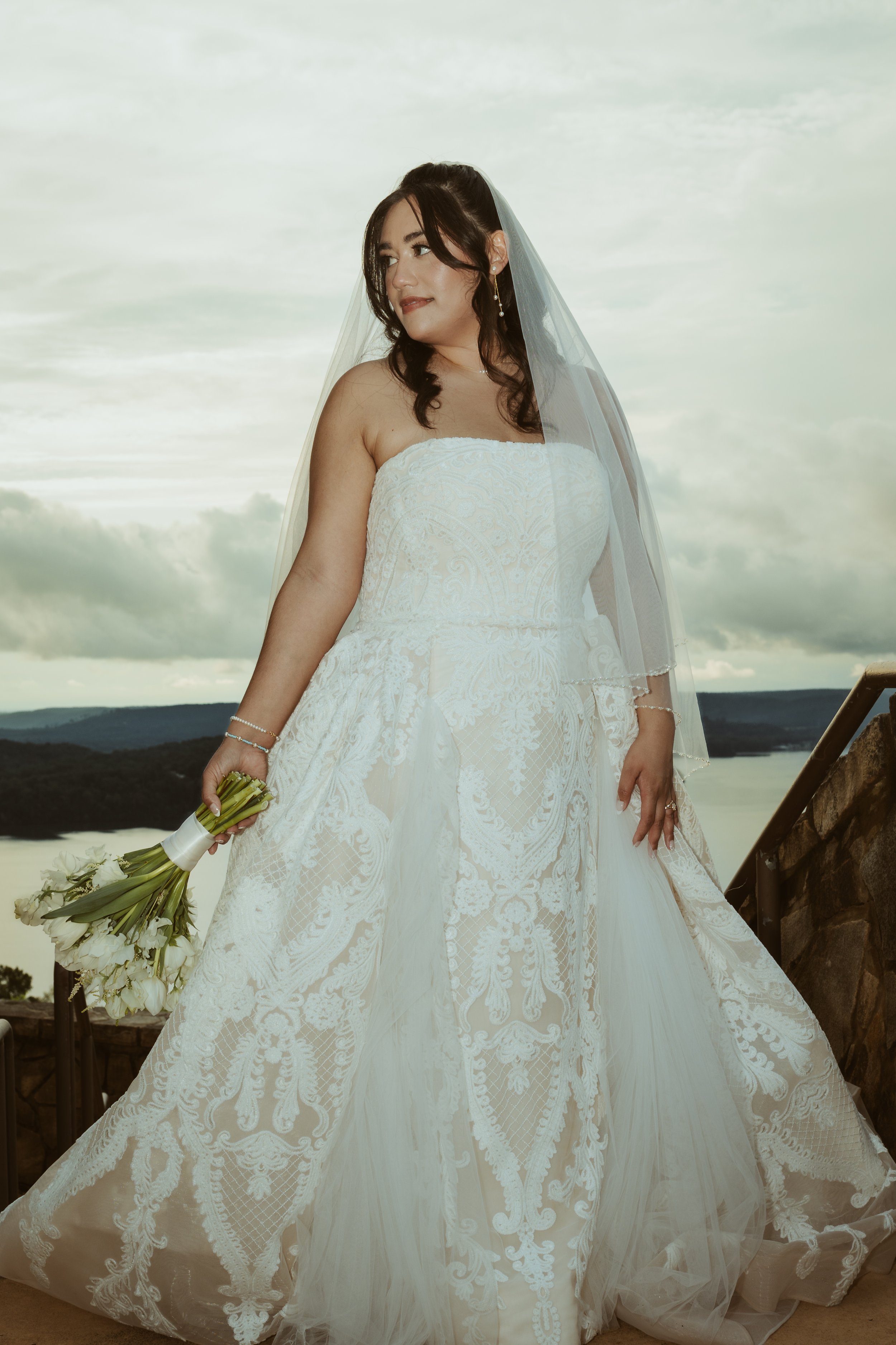A bride in a strapless white gown holding a bouquet of white flowers, standing outdoors with a scenic background of water and hills, cloudy sky.