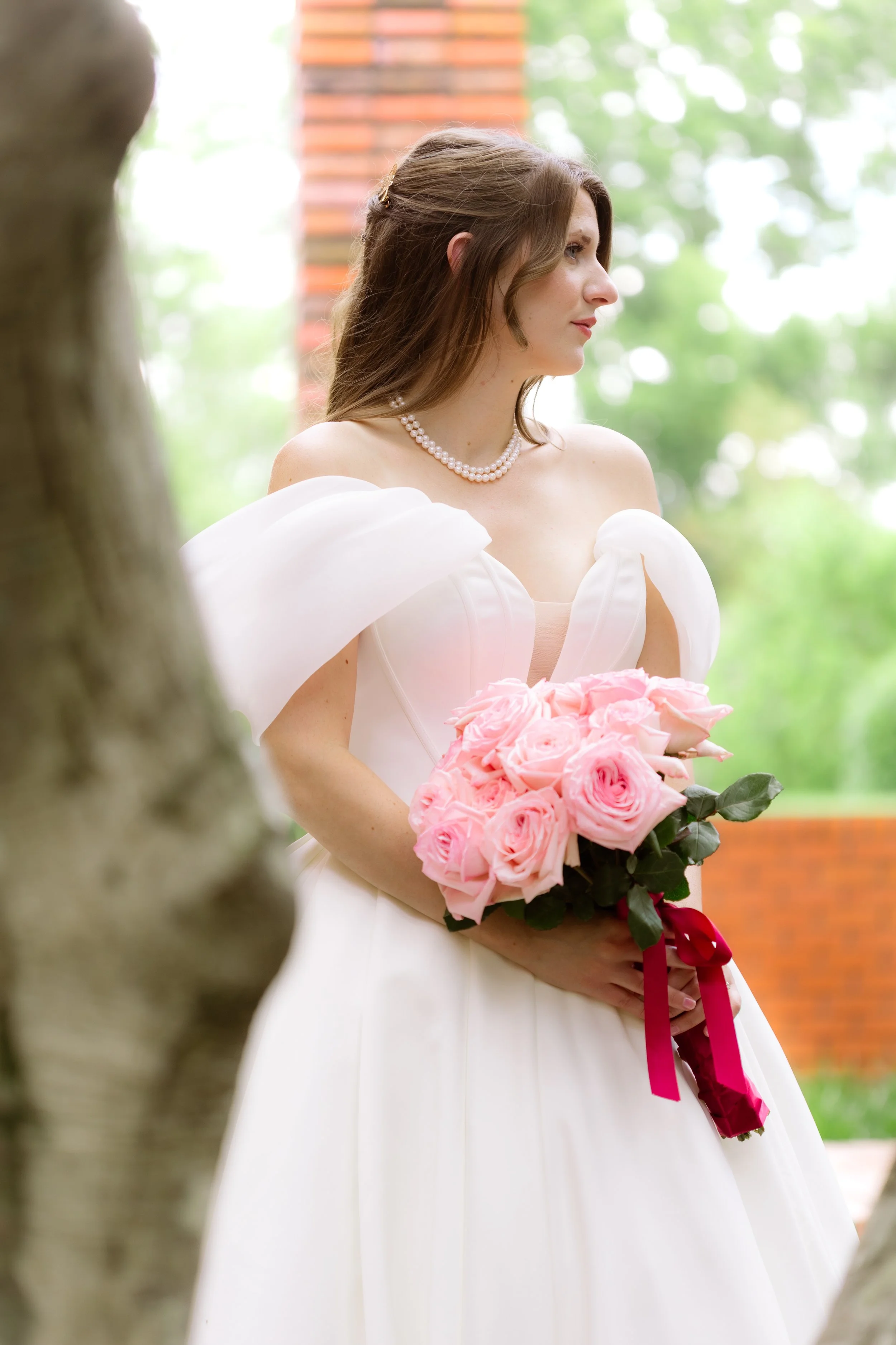 A bride in a white wedding gown holding a bouquet of pink roses. She has brown hair, wears a pearl necklace, and is standing outdoors with green trees in the background.