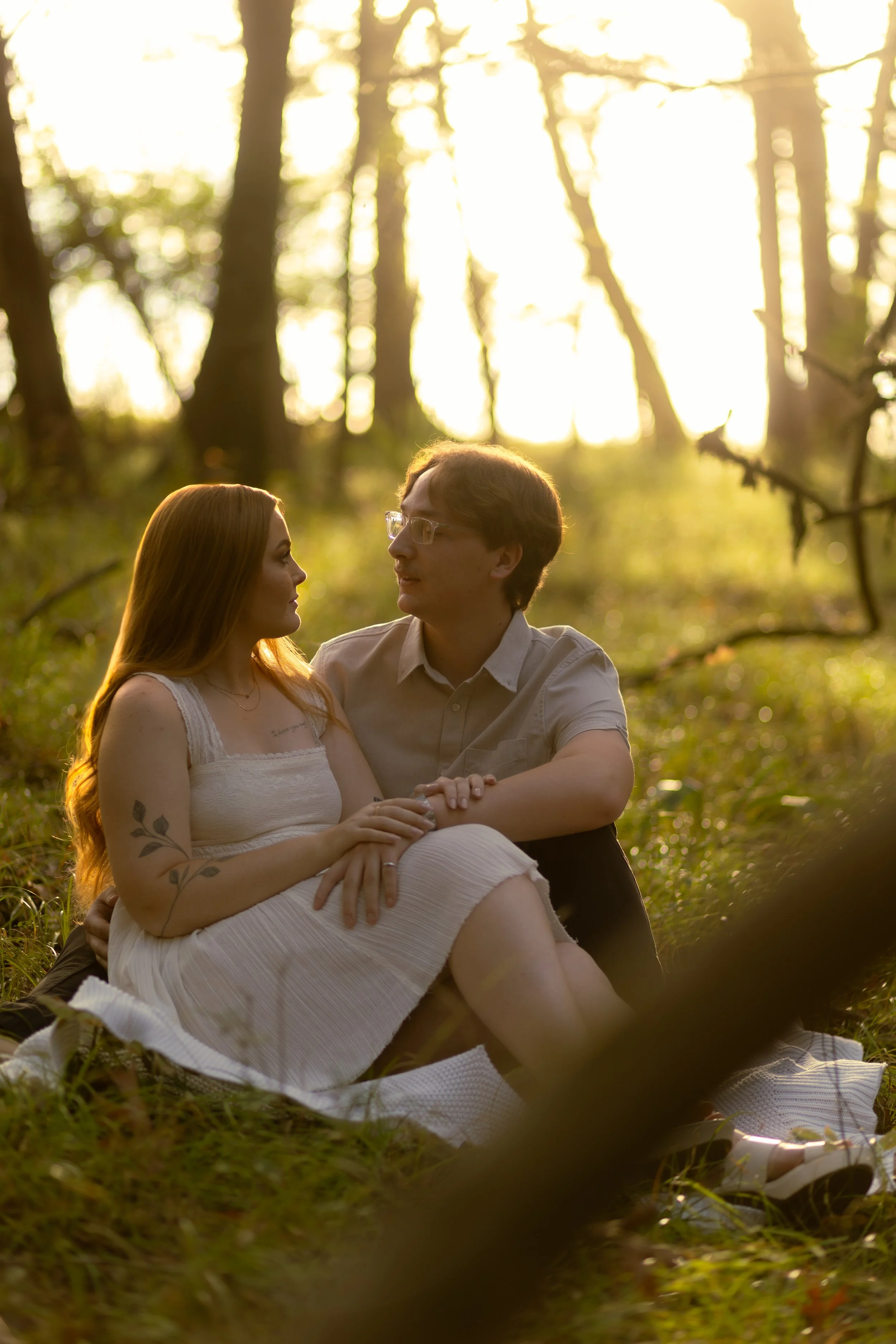 A couple sitting closely together on a blanket in a wooded area during sunset, gazing into each other's eyes.