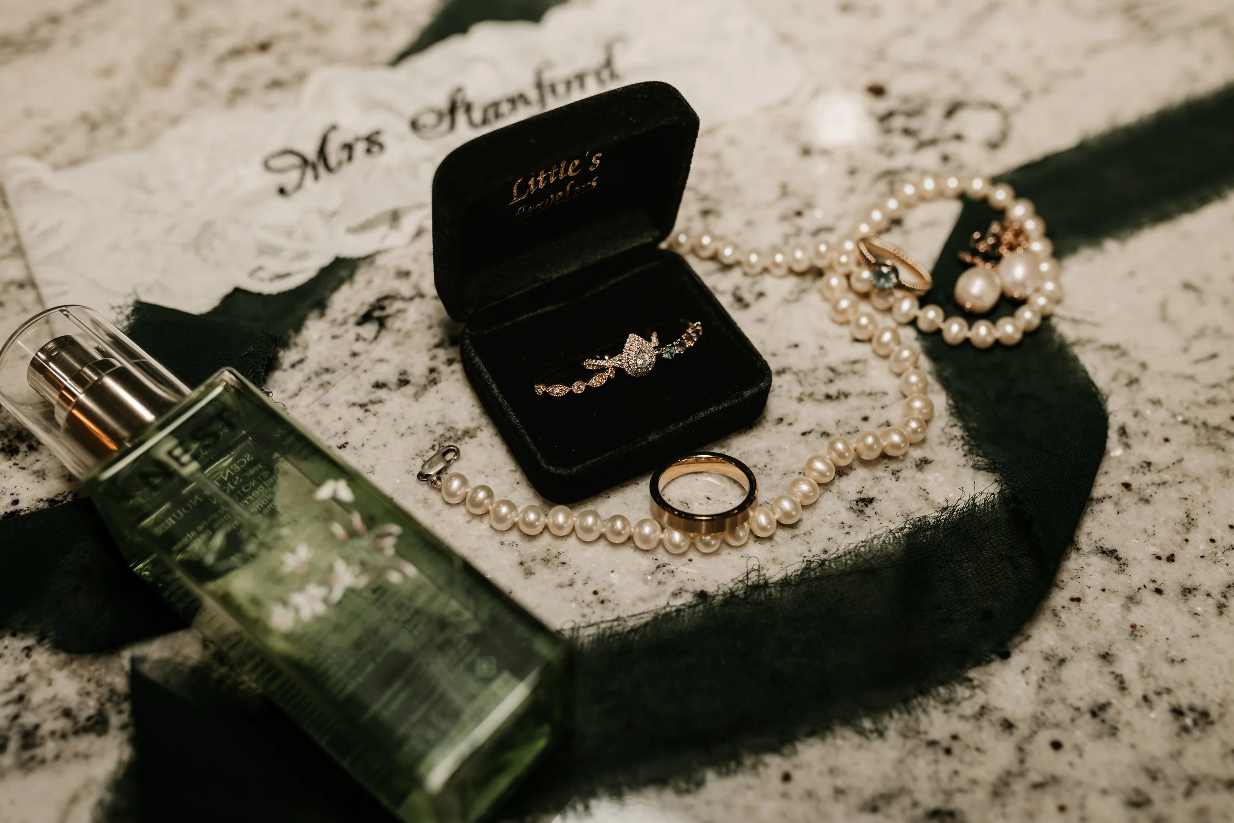 A collection of jewelry and personal items on a granite countertop, including a pearl necklace, a gold ring, a bracelet, a ring box with a diamond ring, a perfume bottle, and a black ribbon.