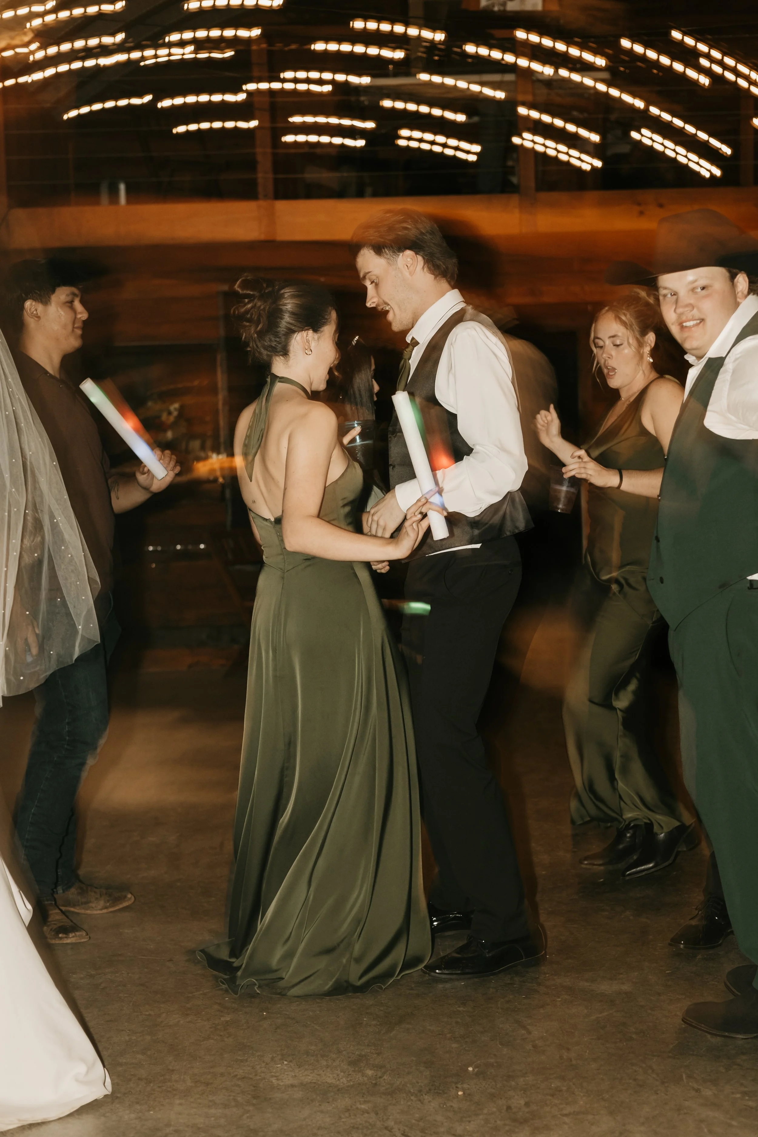 People dancing at a party, holding glow sticks, in a dimly lit venue with wooden decor. Editorial Style wedding reception images in North Alabama