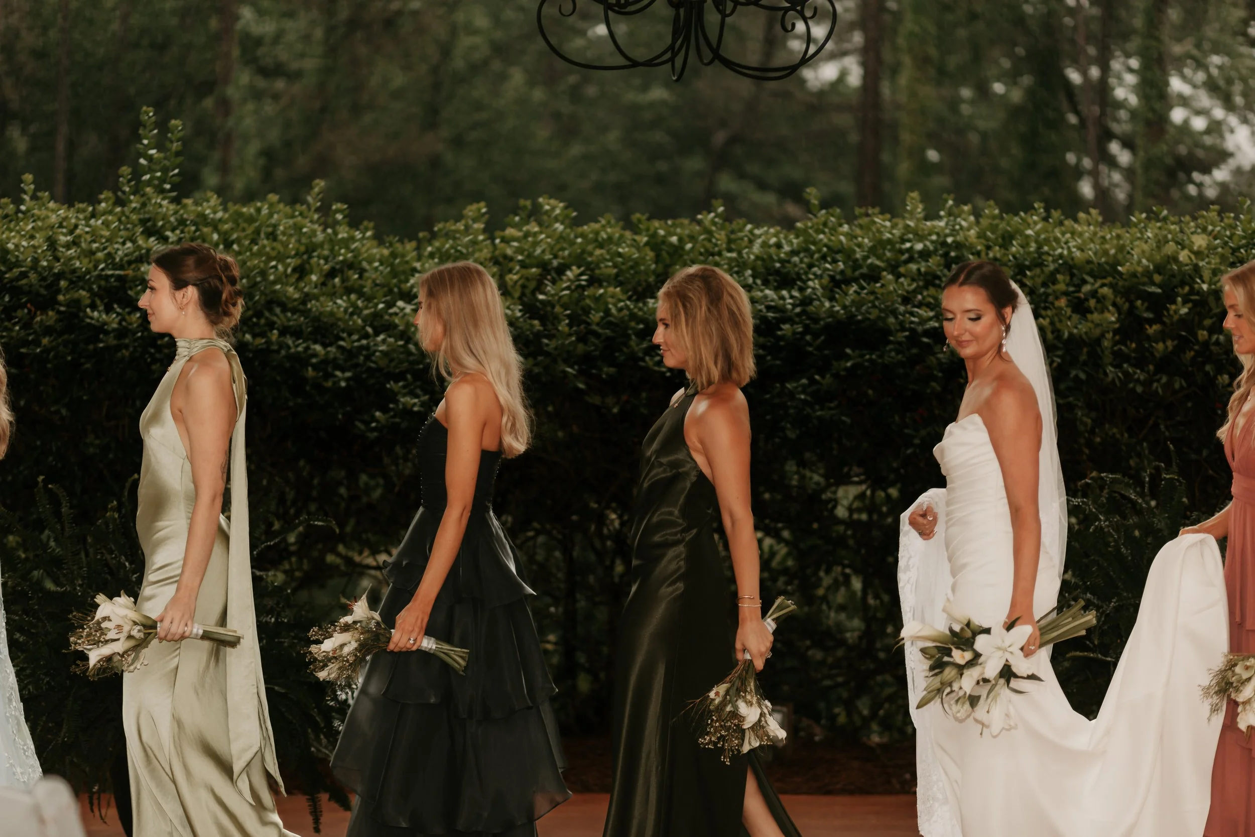 Line of women in formal dresses holding bouquets, walking outdoors on a wedding aisle.