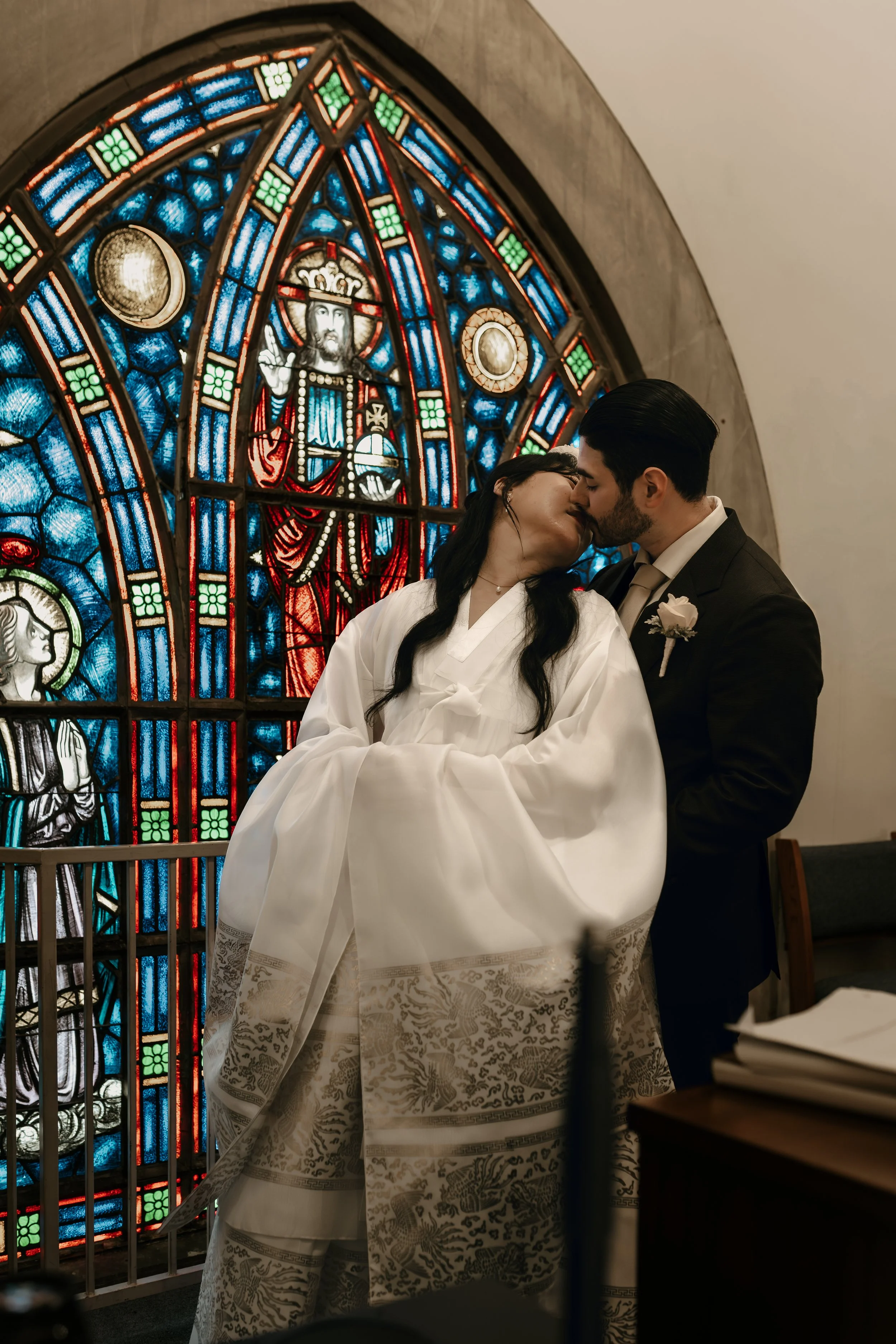 A bride and groom sharing a kiss during their wedding ceremony in front of a colorful stained glass window featuring religious imagery.