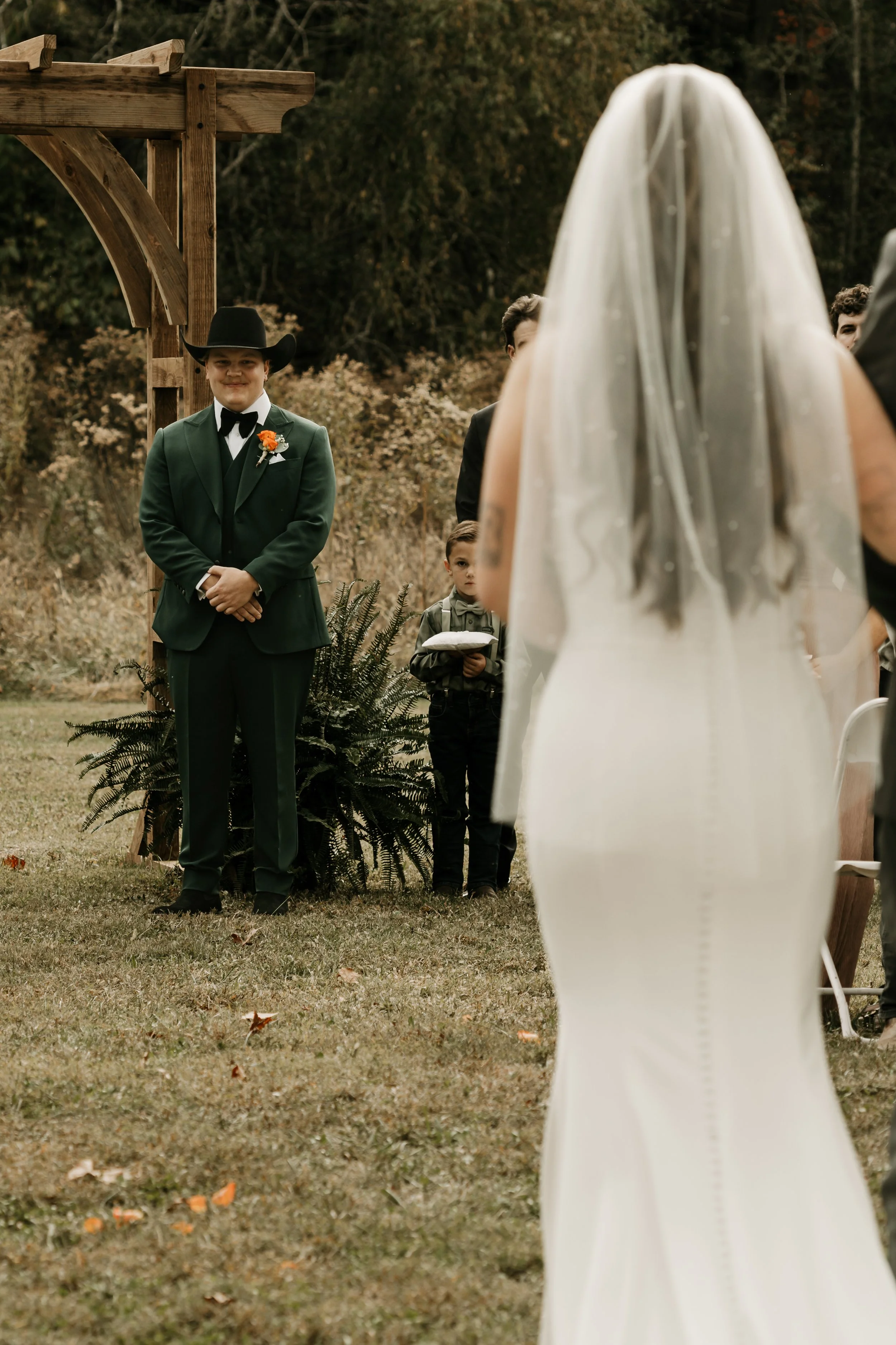 A bride in a white wedding dress and veil facing an officiant at an outdoor wedding ceremony, with a groom in a green suit and cowboy hat standing under a wooden archway in the background.