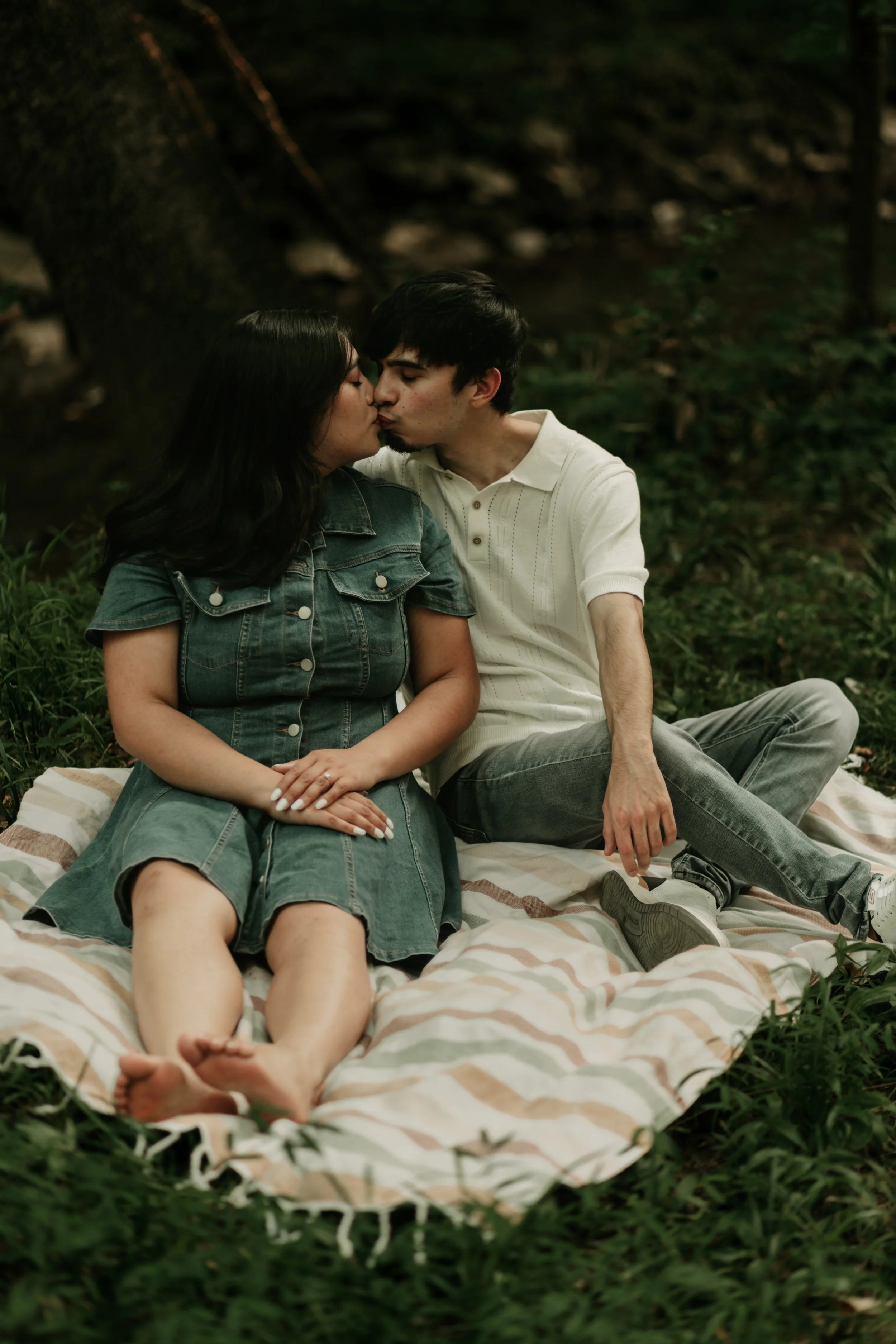 A young couple, sitting on a striped blanket on the grass in a forest, sharing a kiss. Engagement photography in Birmingham, AL.