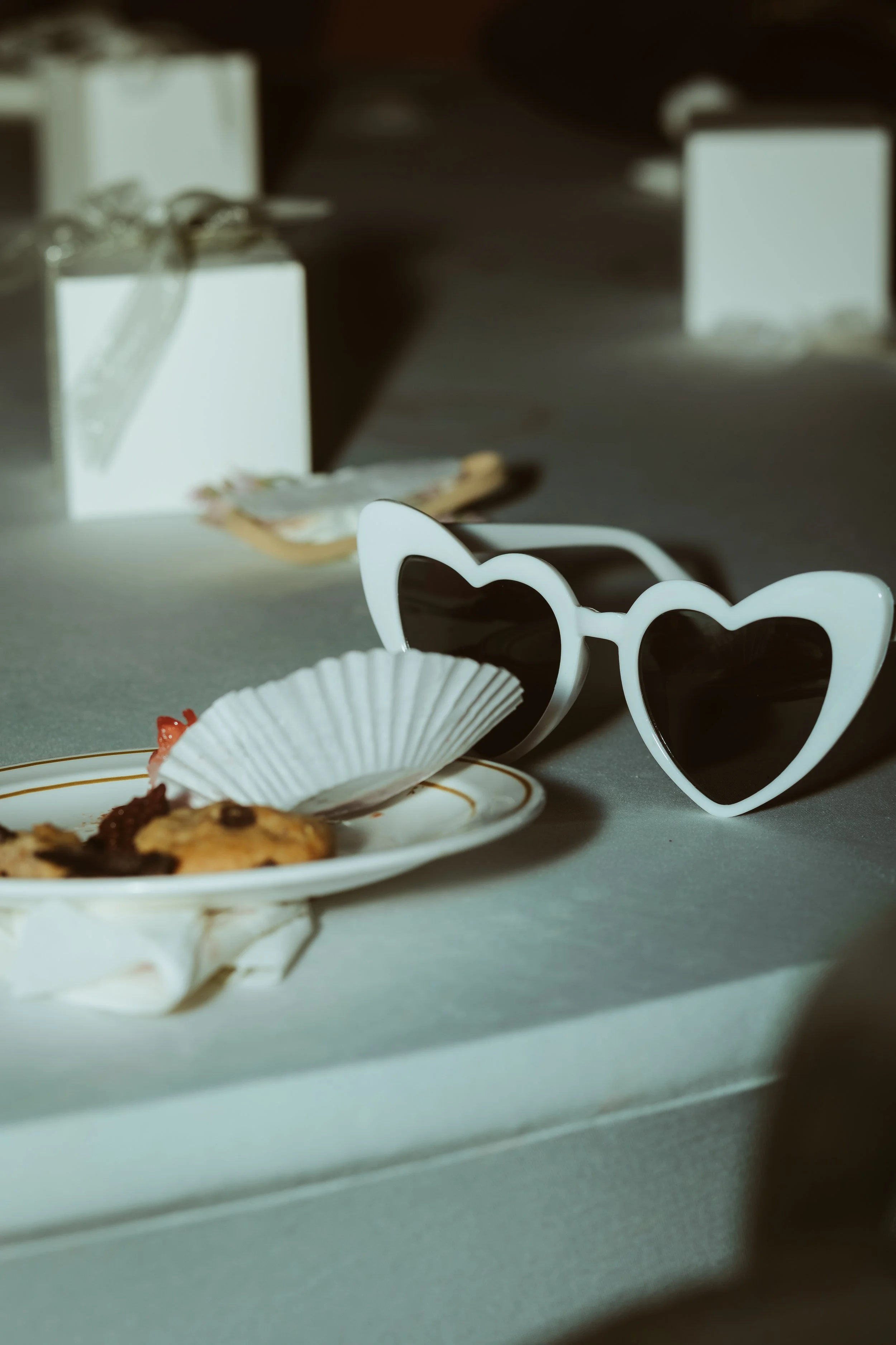 A pair of white heart-shaped sunglasses next to a partially eaten cookie on a decorative white plate, with gift boxes in the background. Fun reception photo ideas.
