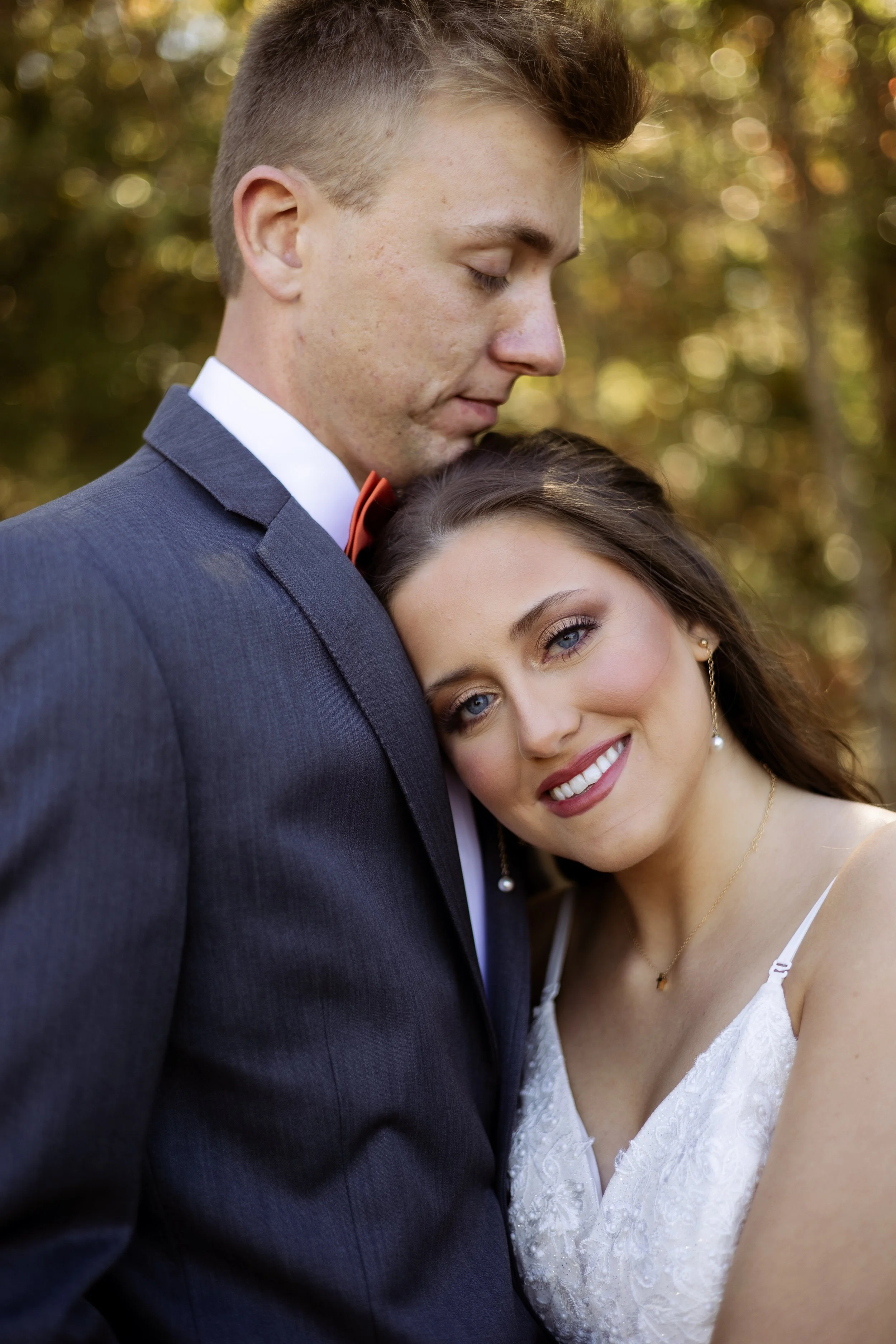 A young couple in wedding attire, the man in a suit with a bow tie and the woman in a white dress, share an intimate moment outdoors during daytime, with blurred trees and sunlight in the background.
