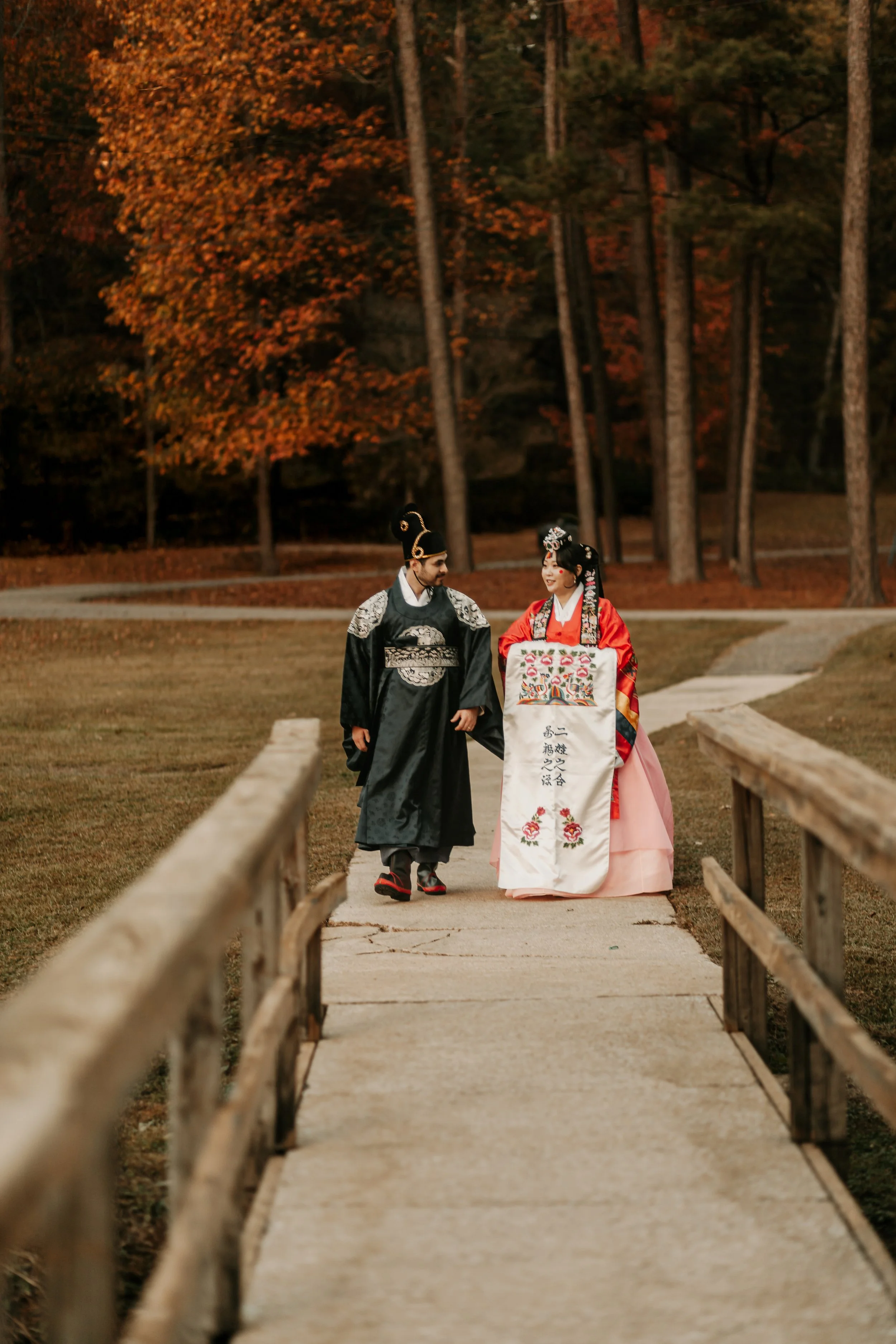 A man and woman dressed in traditional Asian (possibly Chinese or Korean) wedding attire walking on a small bridge in a park with trees showcasing fall foliage.