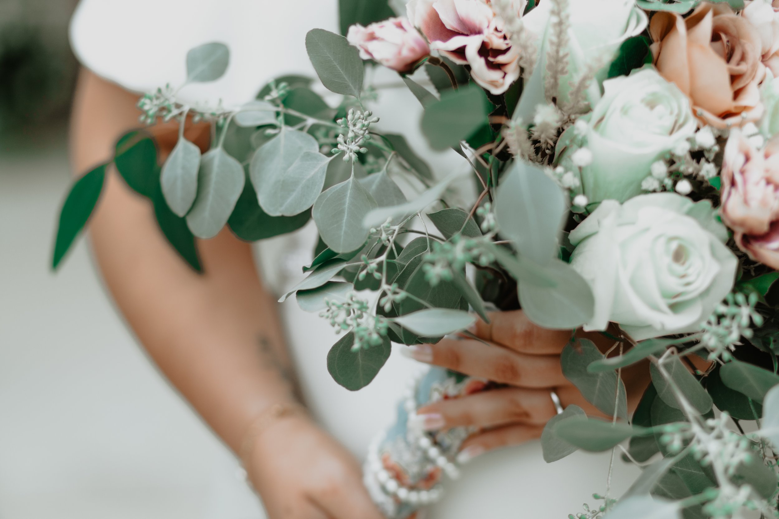 Close-up of a woman holding a bouquet of white roses, pink carnations, and green eucalyptus leaves, with tattooed arm and white beaded bracelet visible