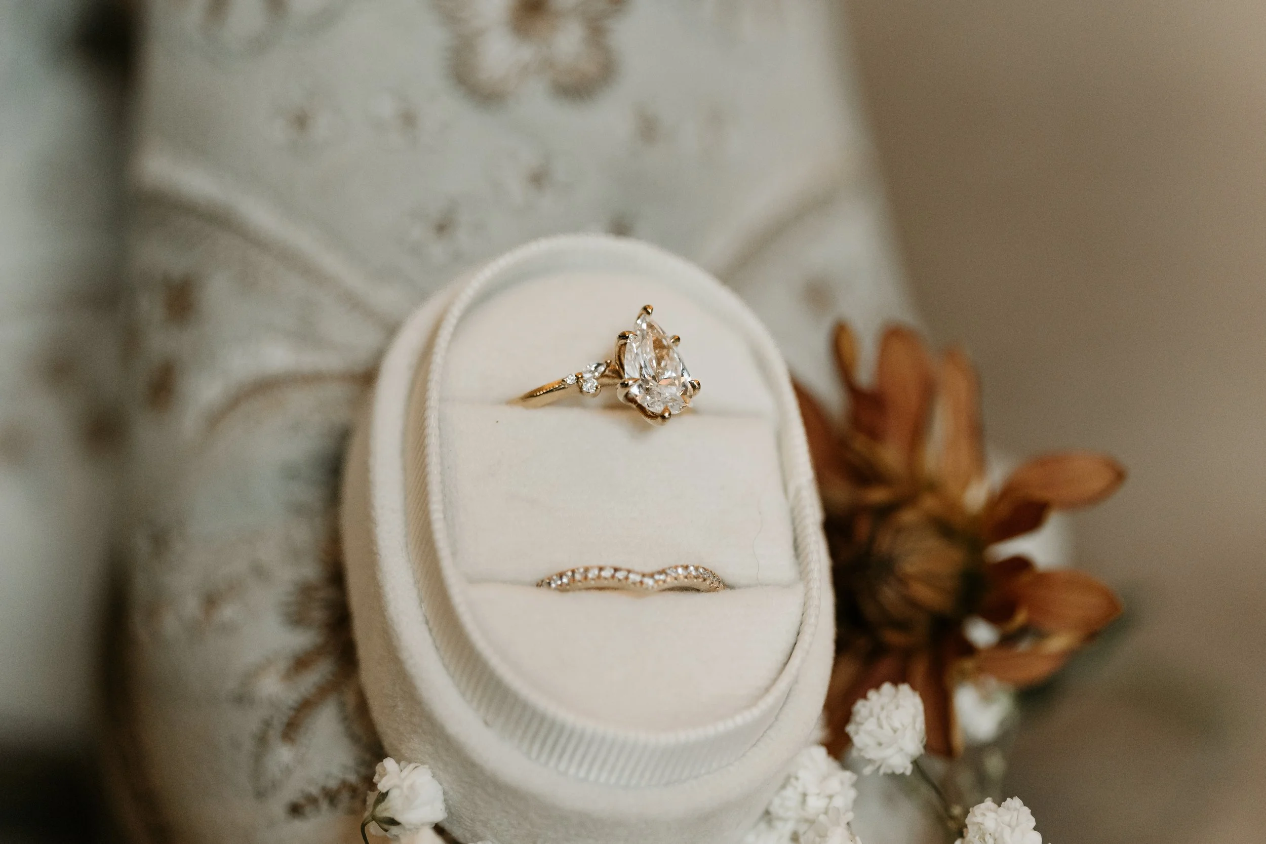 Close-up of a jewelry ring set in an open white velvet jewelry box, with a floral arrangement in the background.