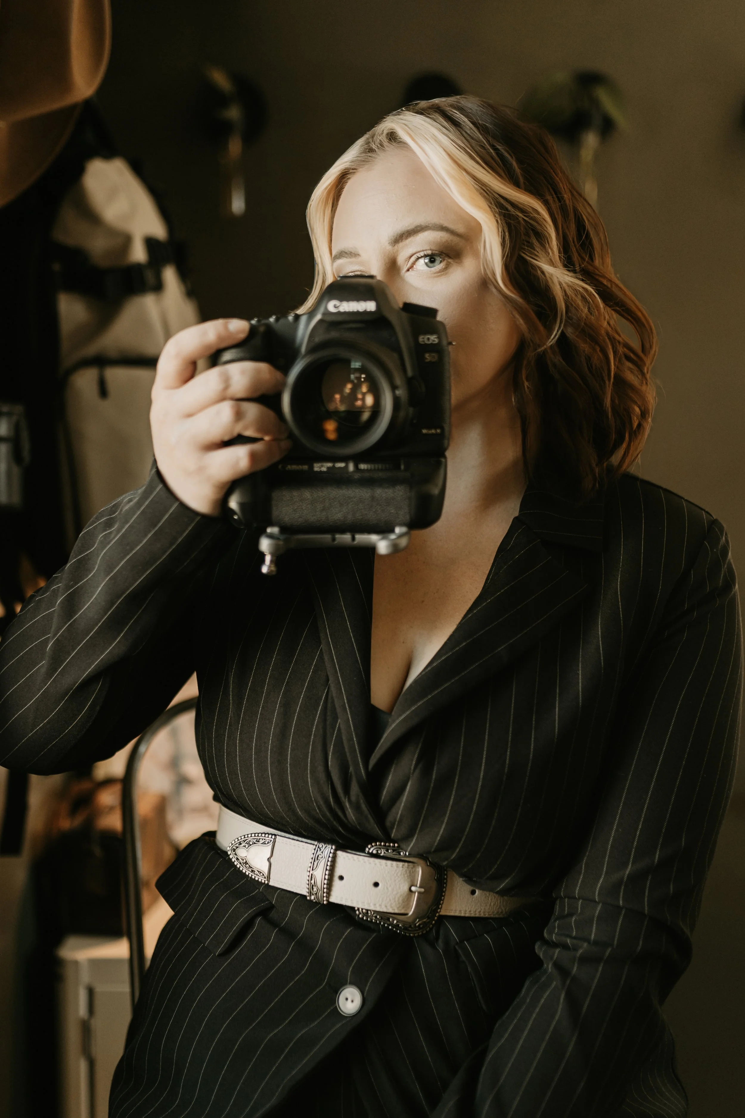 Woman with wavy red hair taking a selfie with a professional camera, dressed in a black pinstripe suit and white belt in a photography studio. Photographer Business Images