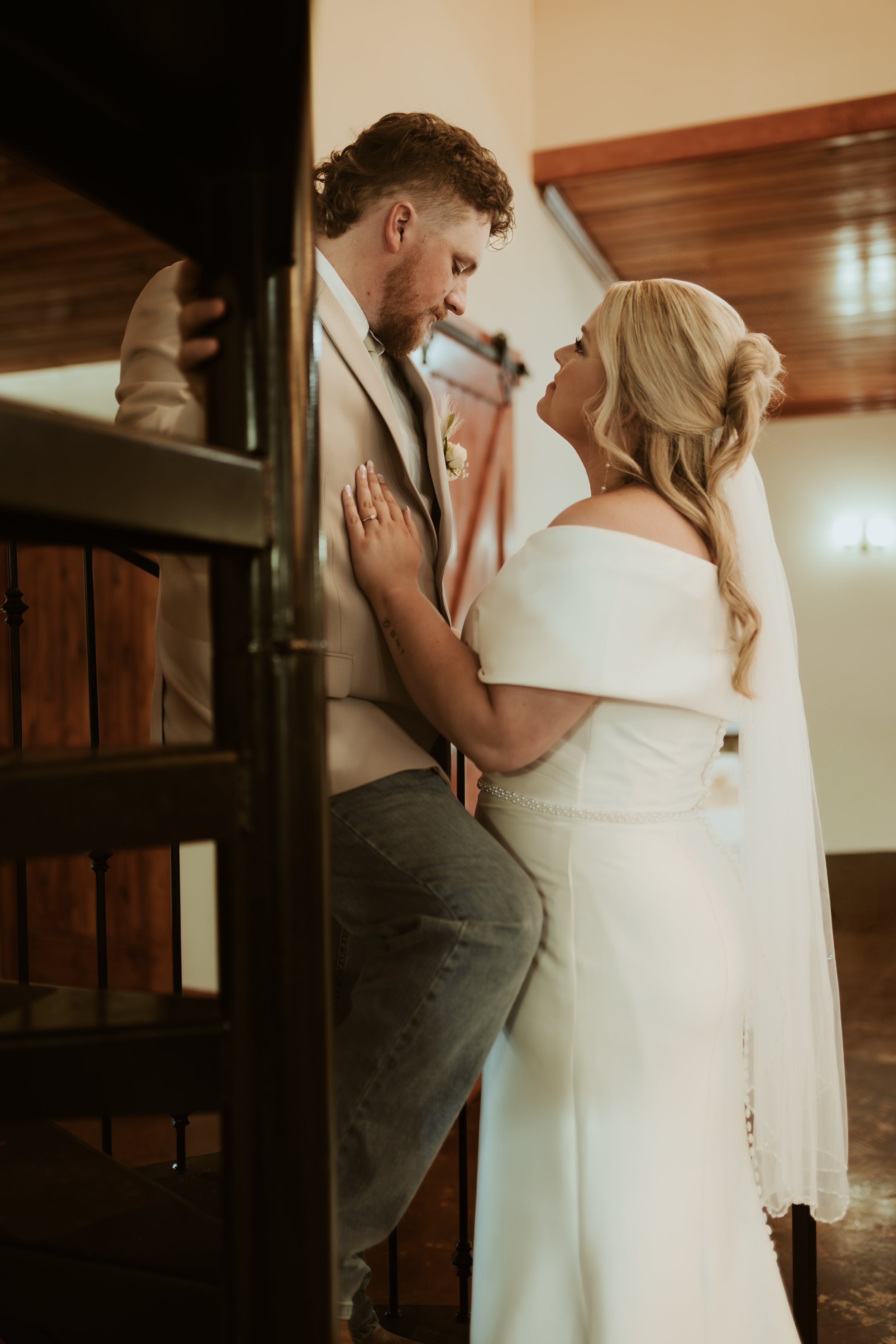 A bride and groom share an intimate moment during their wedding, with the groom sitting on a staircase railing and the bride standing close to him, gazing into each other's eyes. Behind The Pines Wedding Venue in Mississippi.