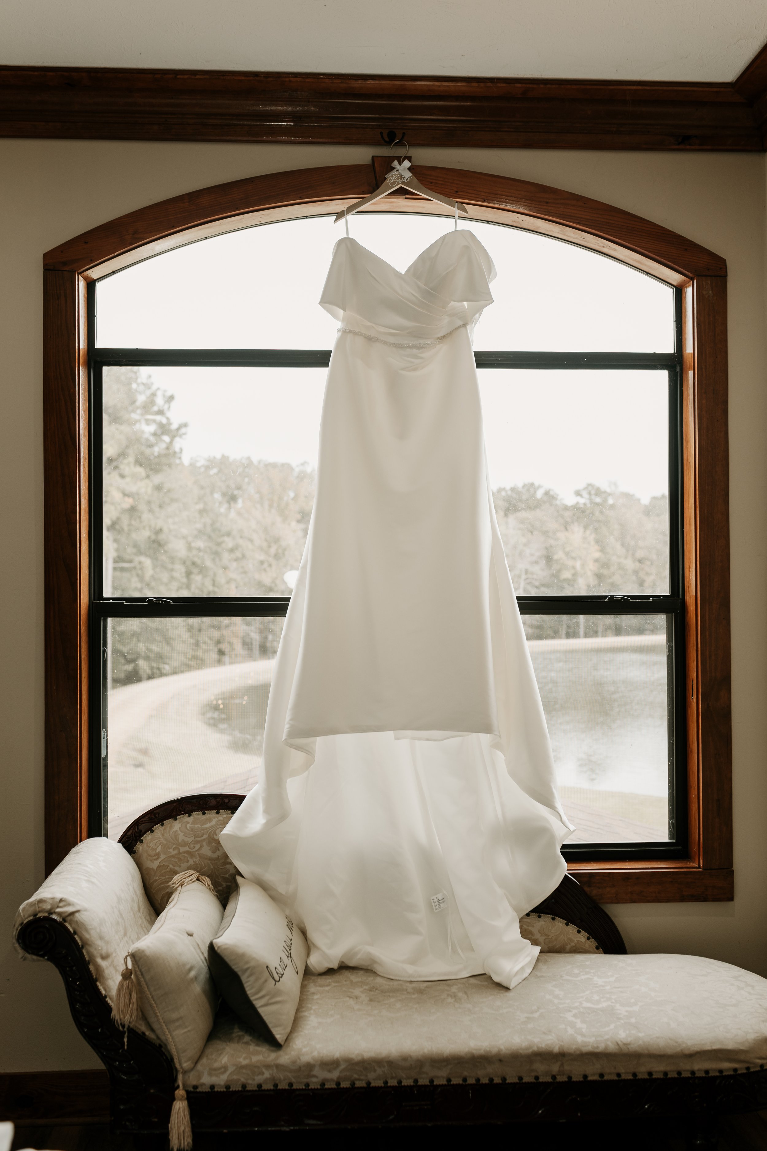 White wedding dress hanging in front of a window with scenic outdoor view. Behind The Pines Wedding Venue and Wedding Photography by Candace Morris with Sixteen Seventeen Photography